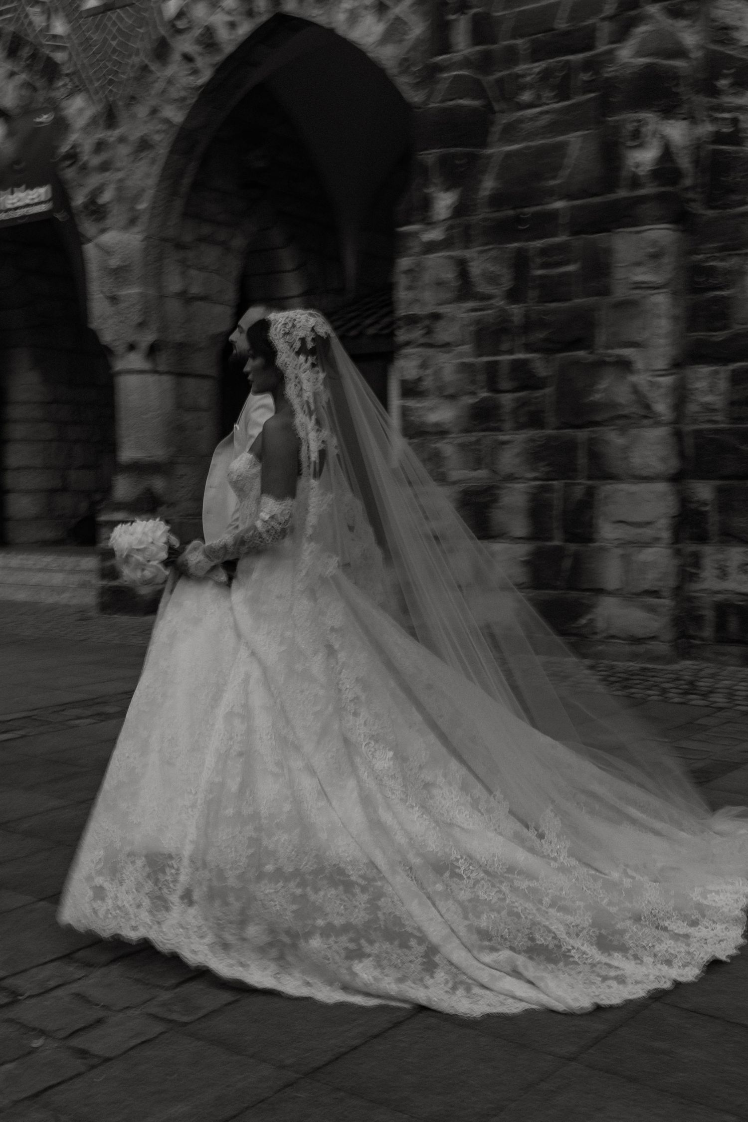 Black and white photo of a bride in lace gown and veil holding bouquet outside a stone church.