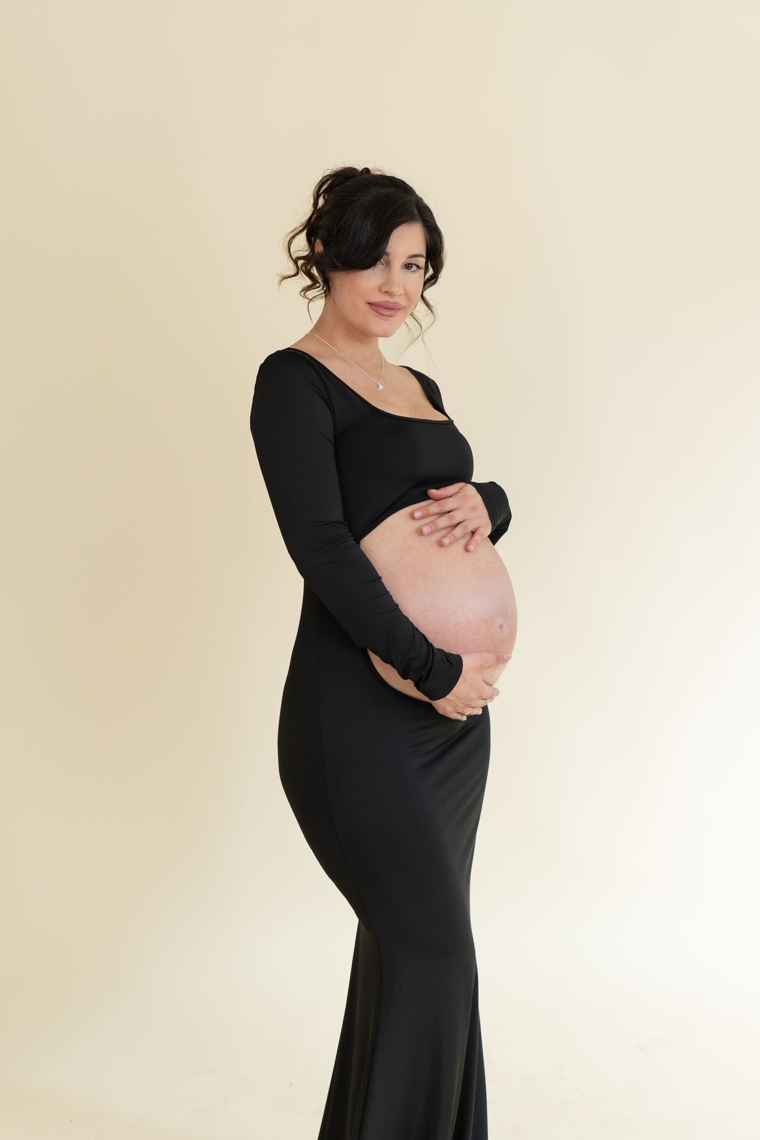 Elegant pregnant woman in black gown cradling her baby bump while posing against a beige background.