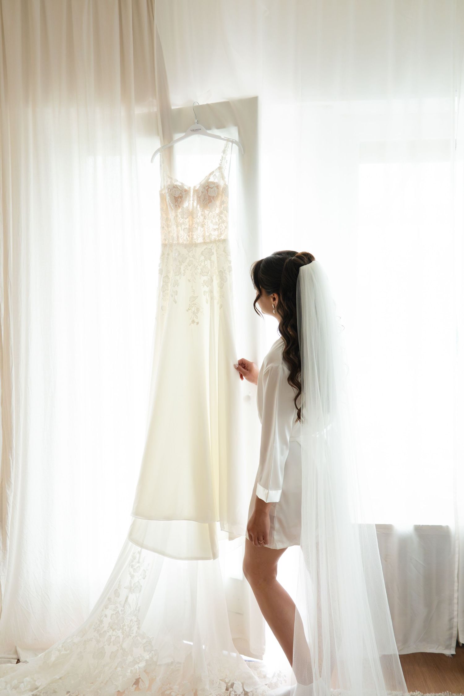 Elegant bride in white robe gazing at her lace wedding gown hanging near a bright window.