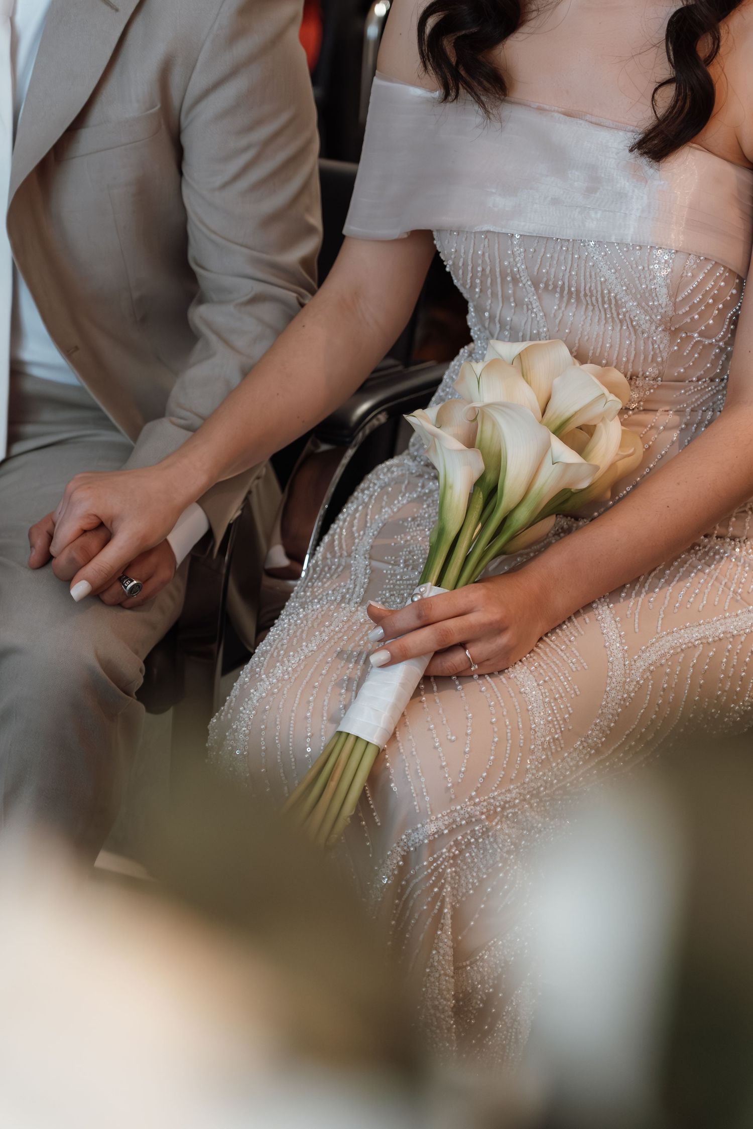 Bride in beaded gown holds white calla lily bouquet while sitting beside groom in tan suit during wedding ceremony.