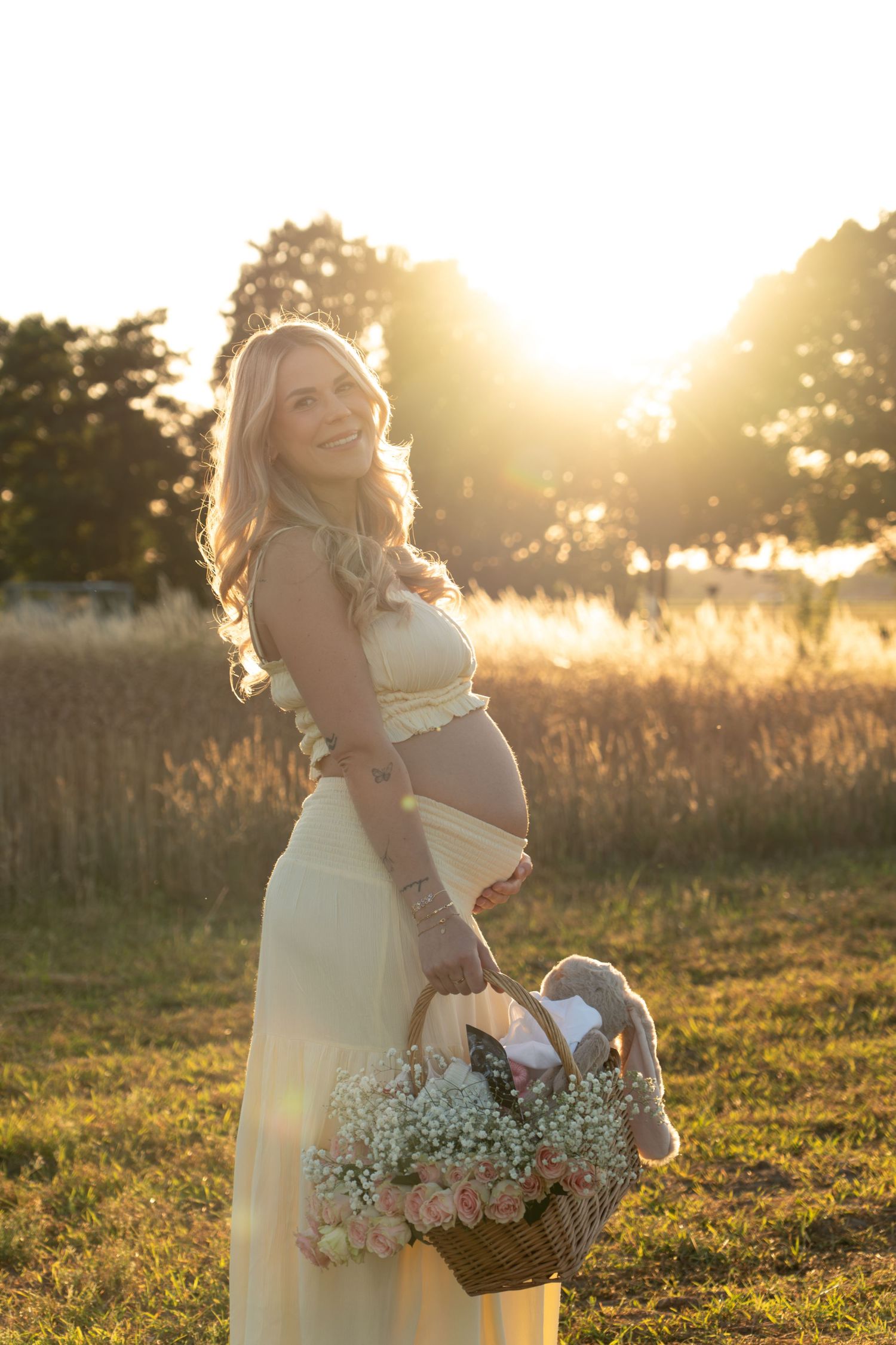 Smiling pregnant woman in yellow dress holding flower basket at golden sunset in field.