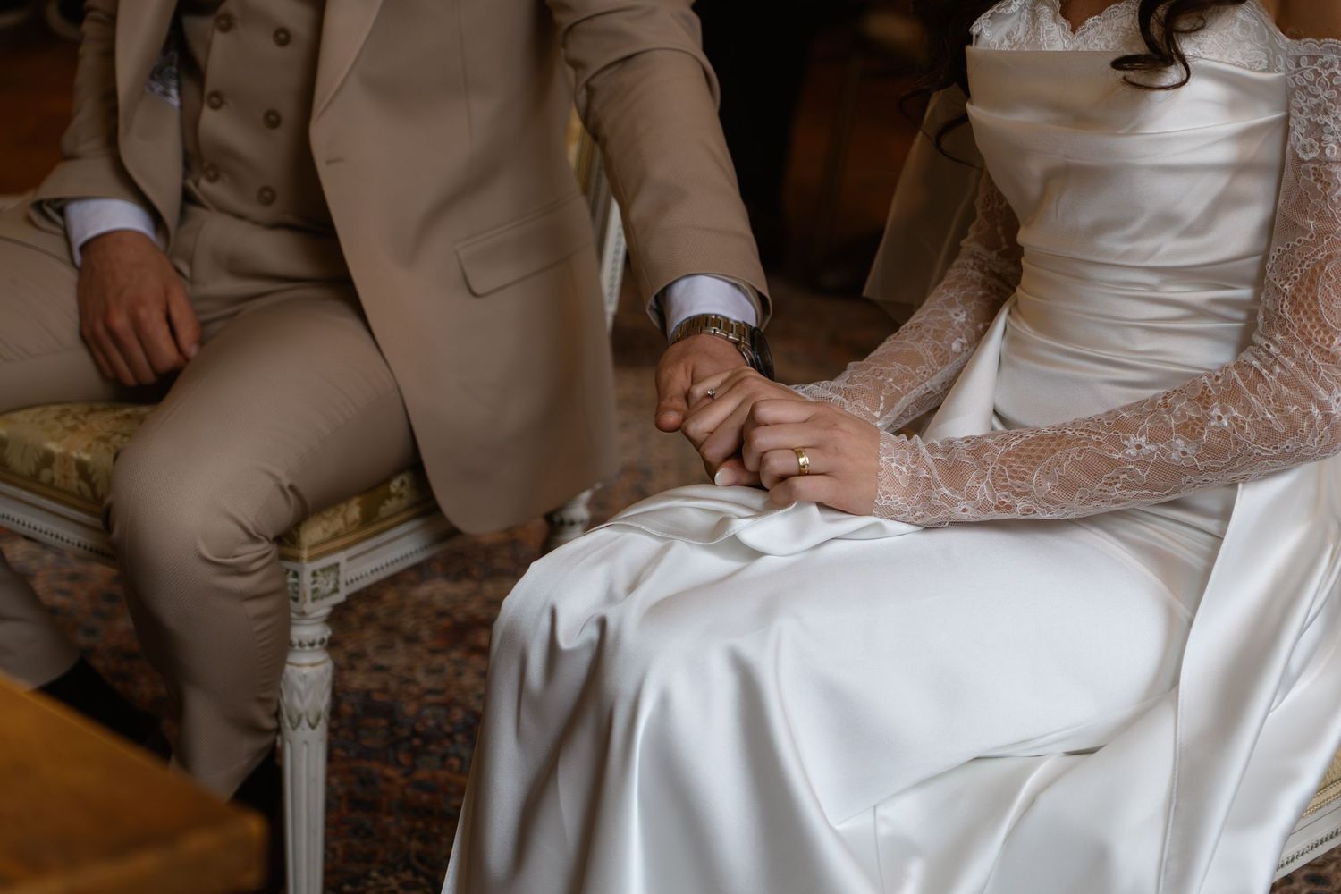 Bride and groom holding hands in wedding attire while seated during ceremony.