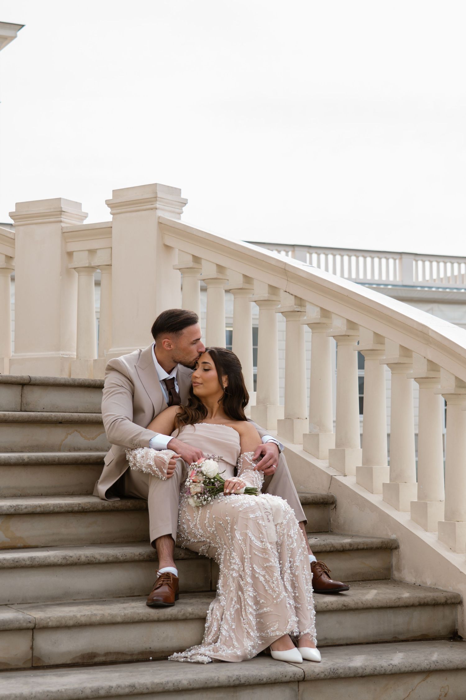 Groom in beige suit kissing bride in embellished gown seated on elegant stone staircase.