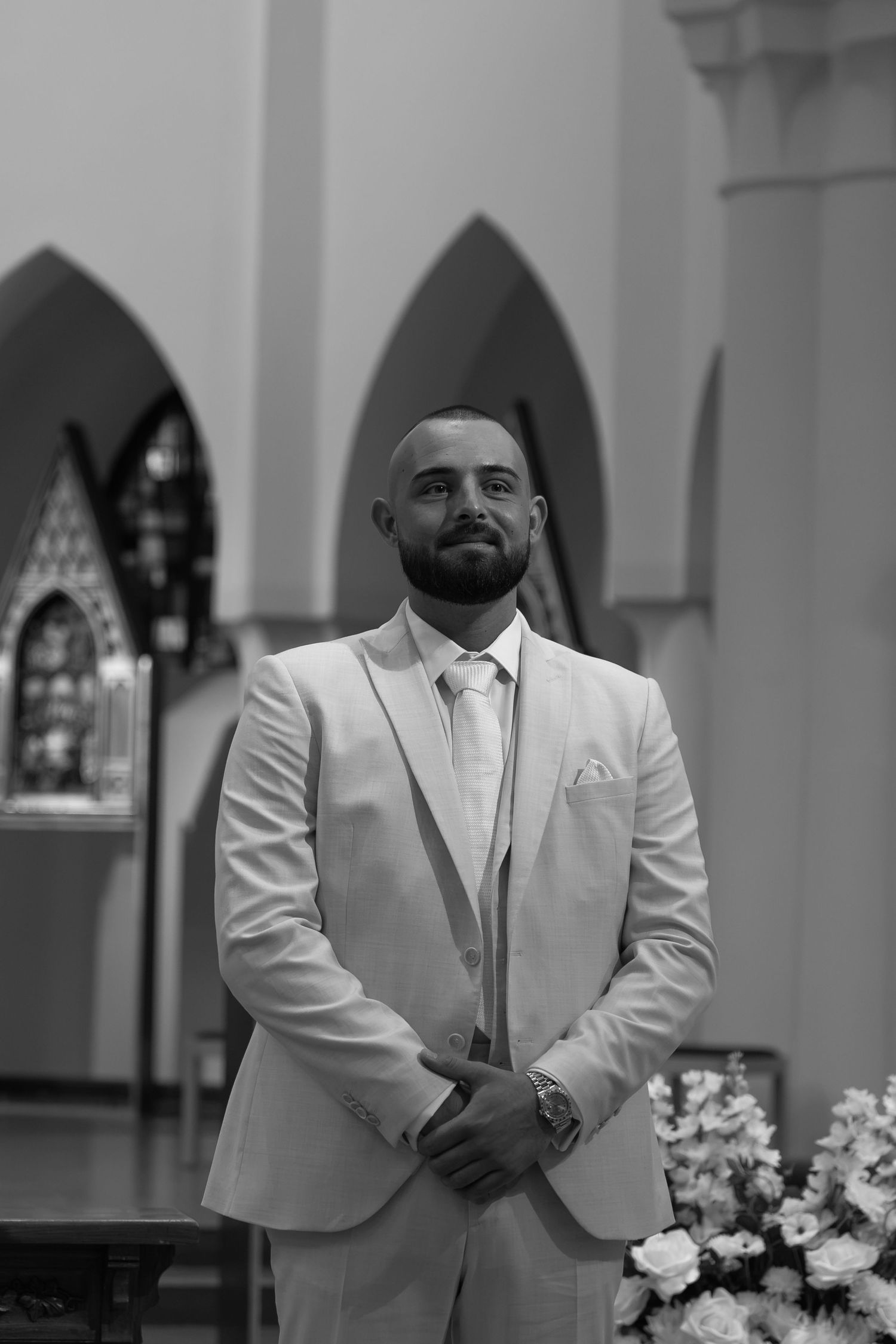 Black and white photo of a bearded groom in a light suit standing in a church, looking upward with hands clasped.