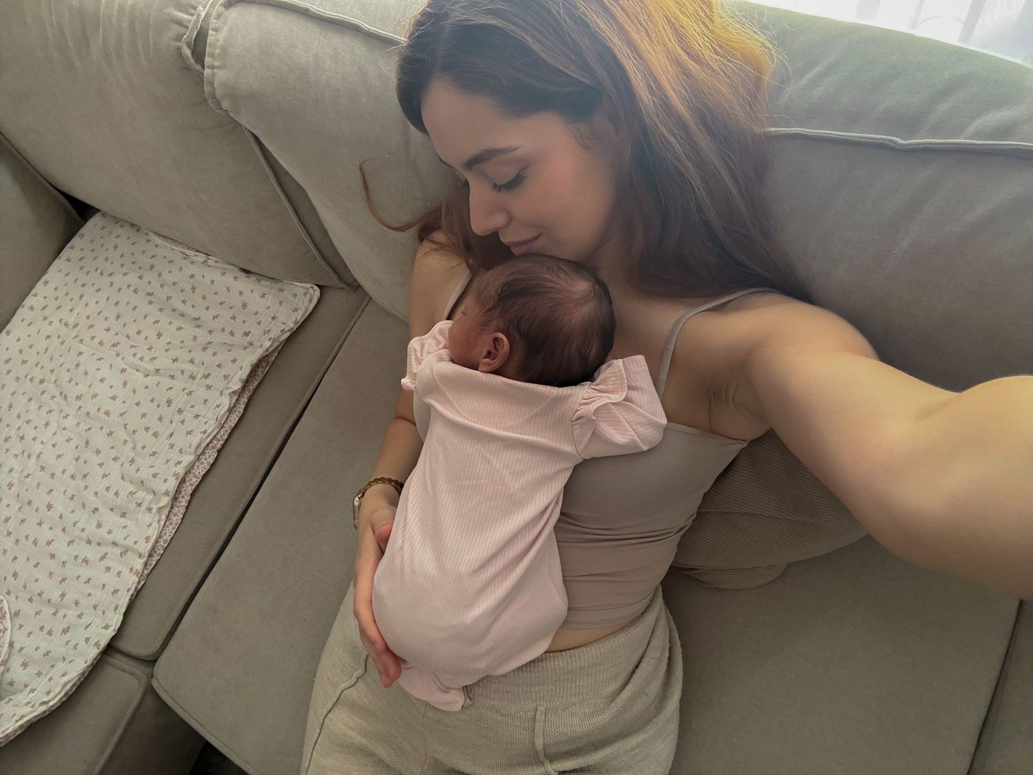 Mother tenderly kissing her newborn baby while resting on a gray sofa in a cozy home setting.