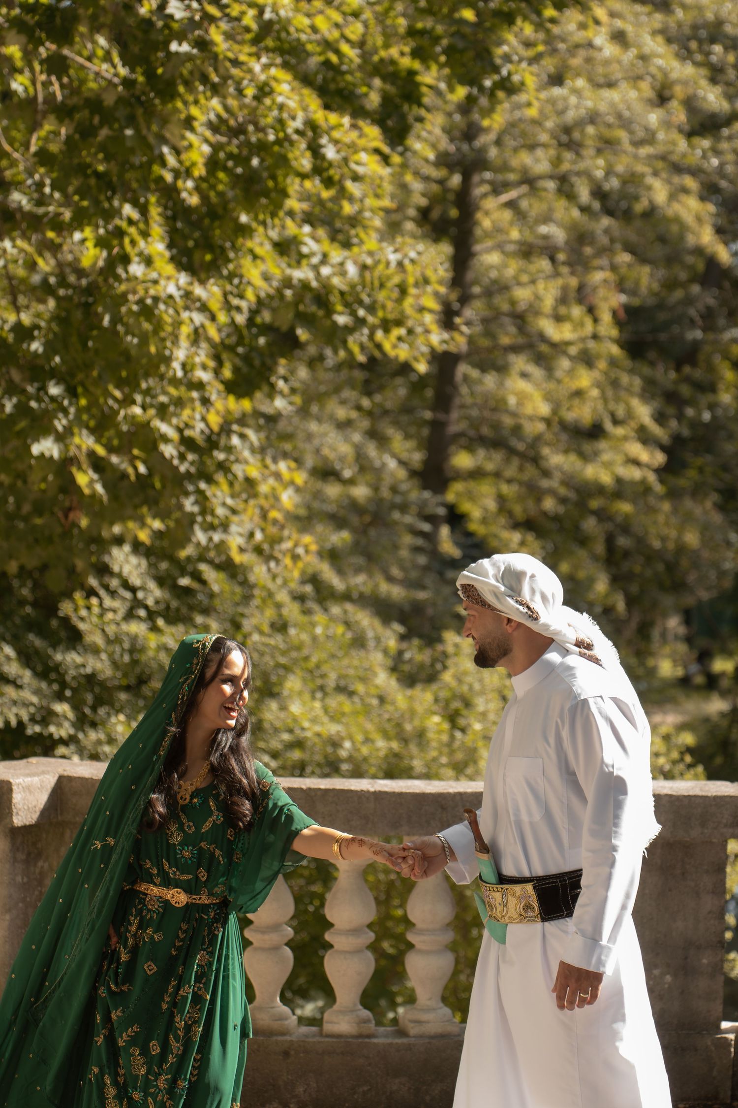 Couple in traditional Middle Eastern attire holding hands on stone balcony surrounded by lush green trees.