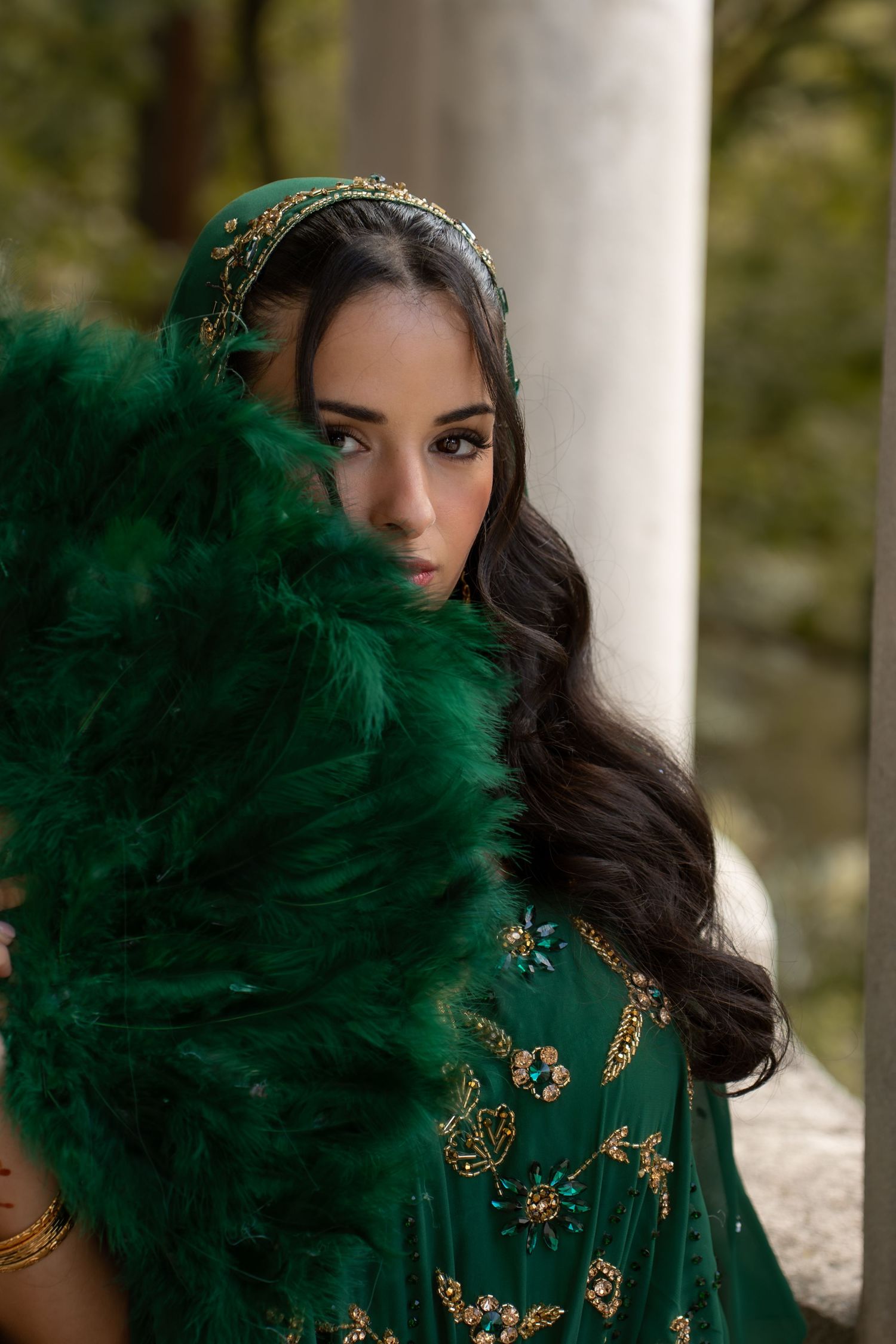 Stunning bride in emerald green embroidered lehenga holds matching feather fan, wearing ornate headpiece among forest columns.