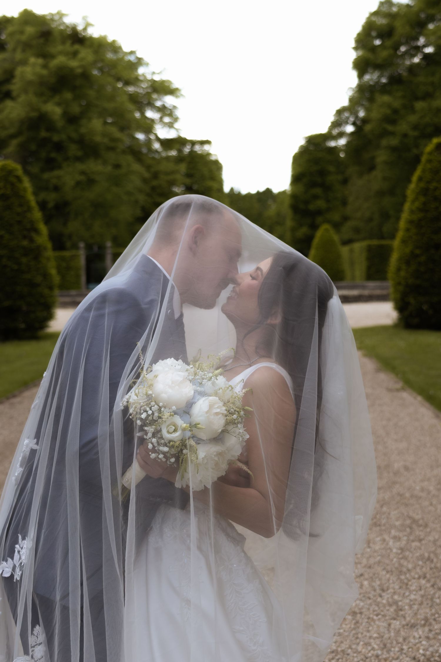 Bride and groom sharing a kiss under a flowing white veil, holding a white floral bouquet outdoors.