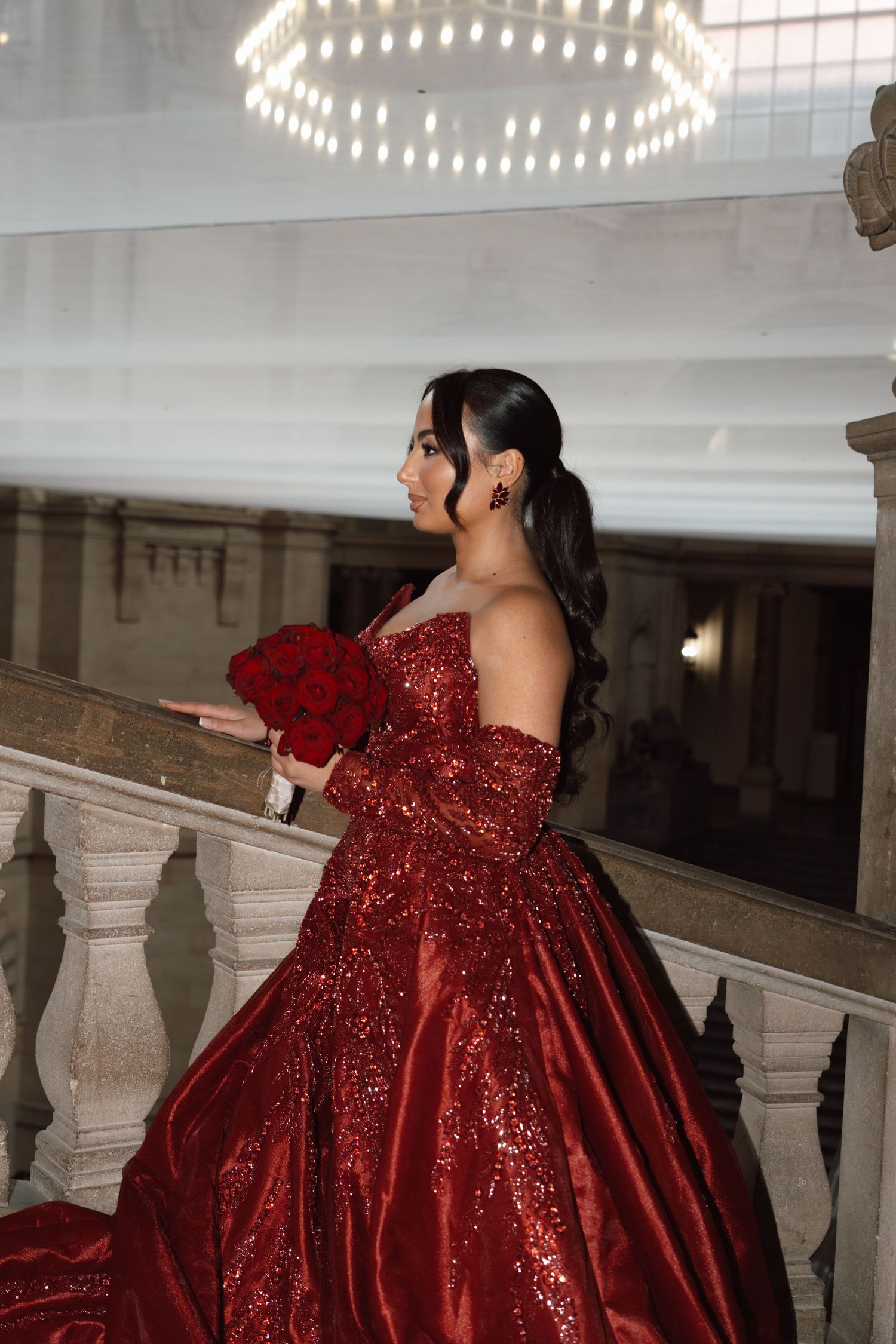 Woman in red sequin ball gown holding red roses bouquet on elegant staircase.