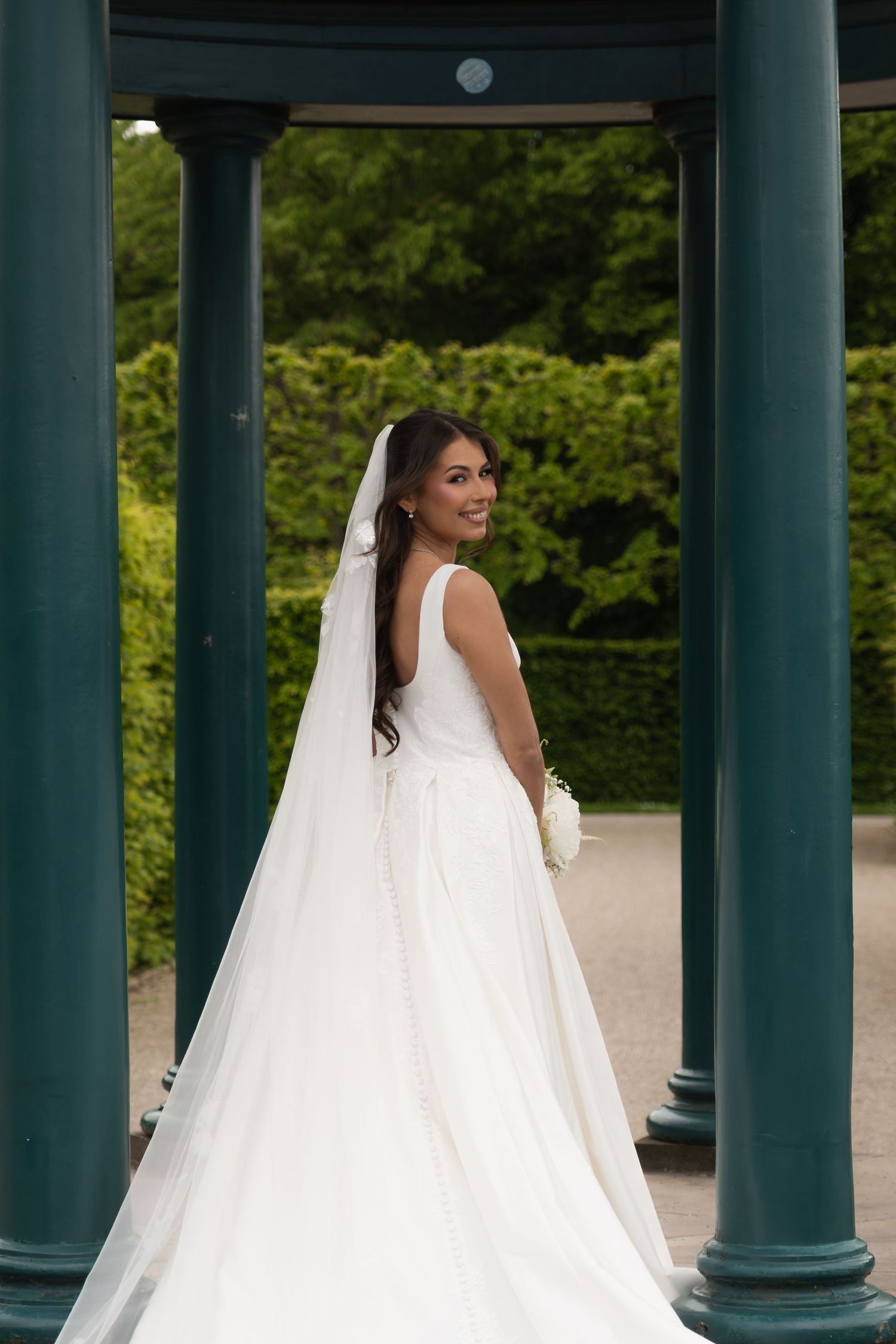 Bride in backless white gown with veil smiling over shoulder between dark green columns.