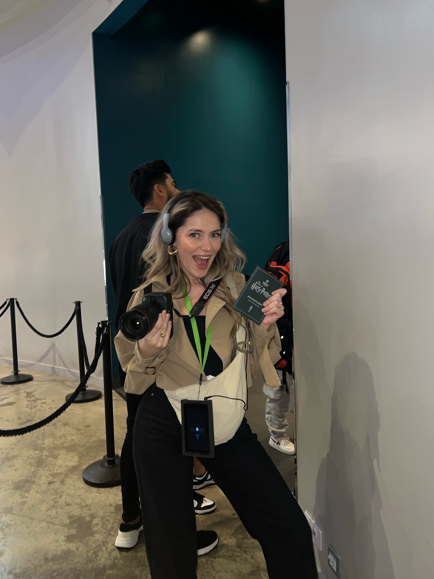 Excited woman with headphones holds camera and badge while waiting in line at event.
