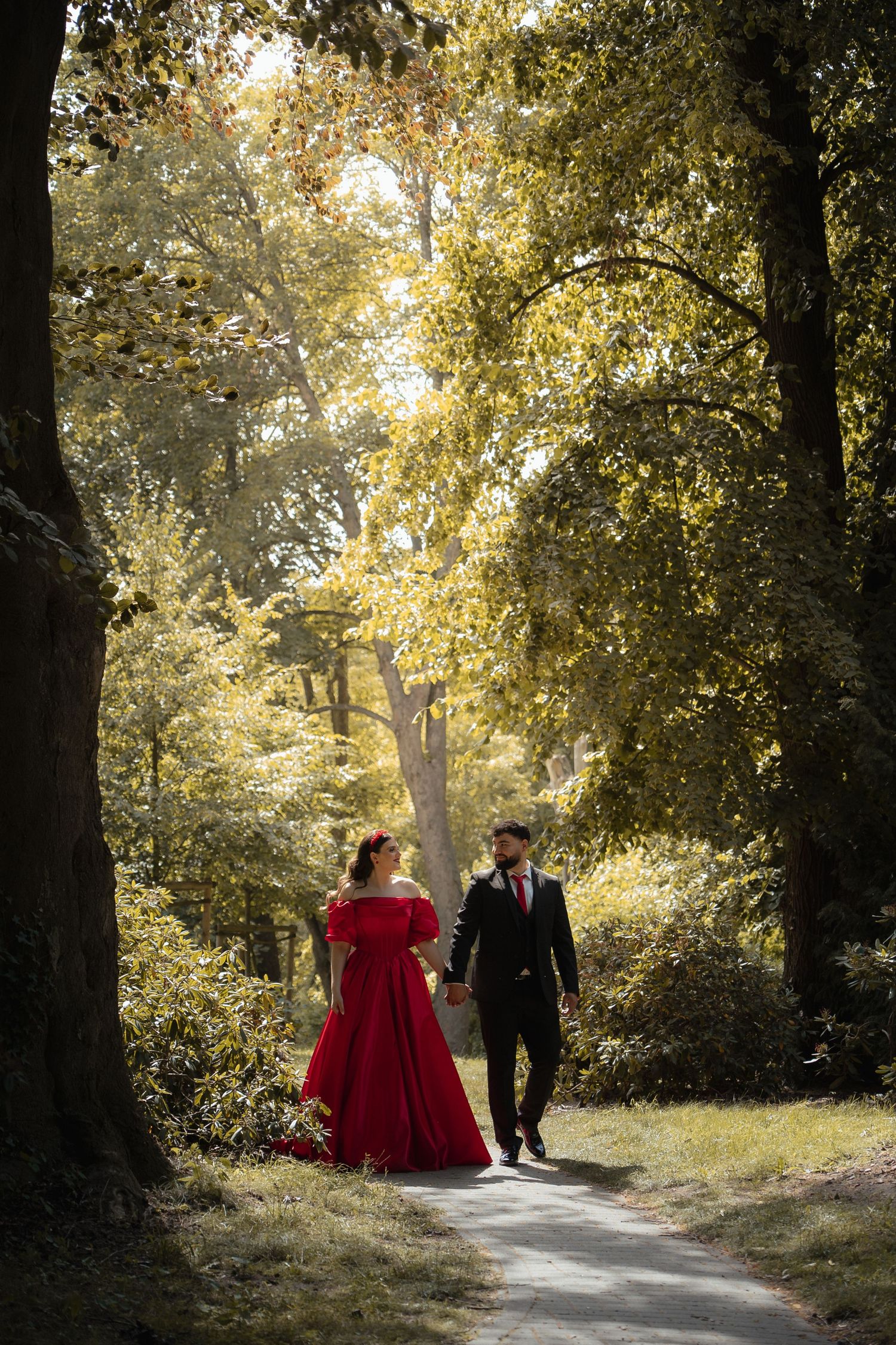 Couple walking hand in hand on a sunlit forest path, bride wearing a stunning red gown.
