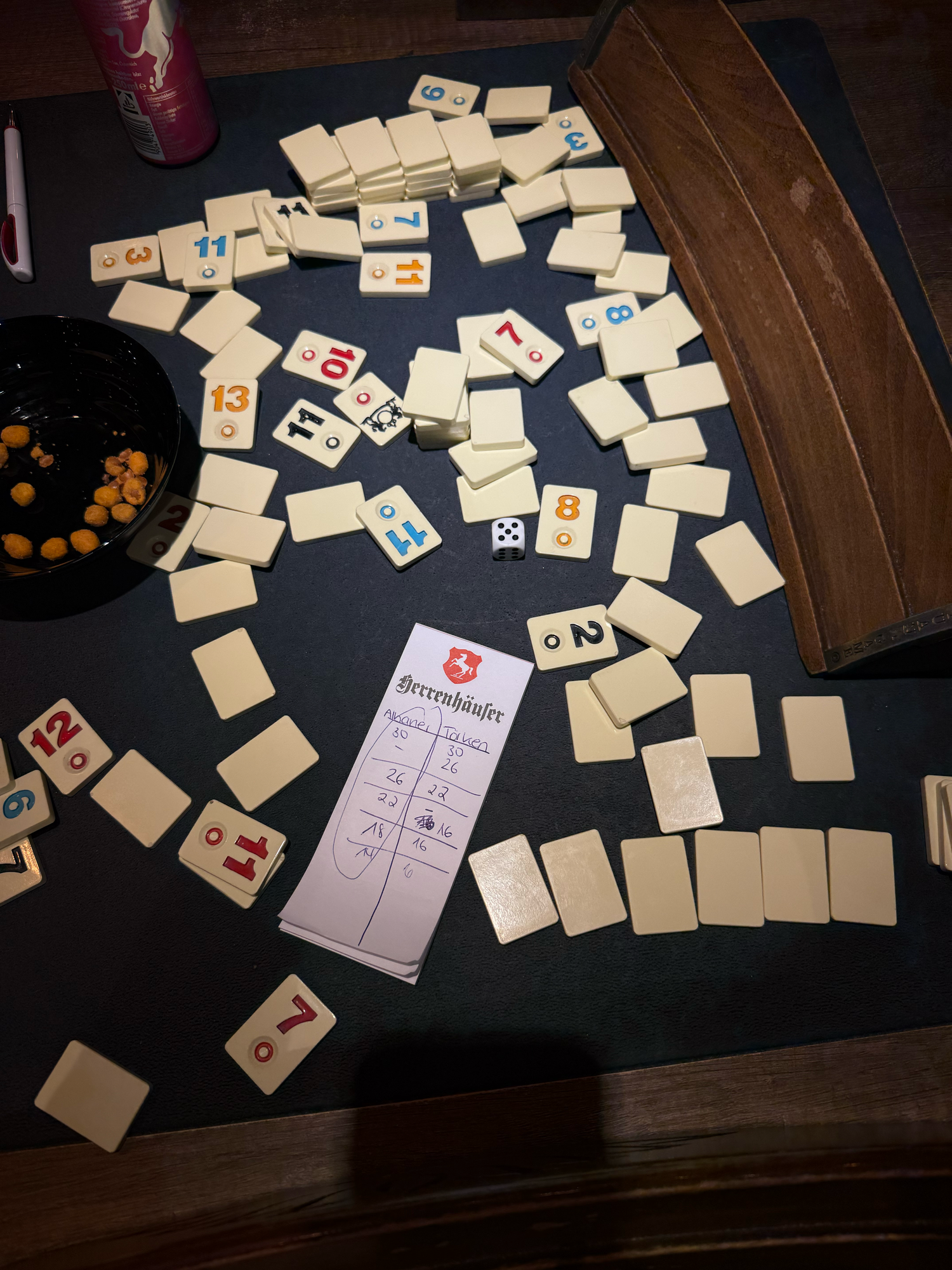 Rummikub tile game spread across a dark table with score sheet, dice, and a bowl of game pieces.