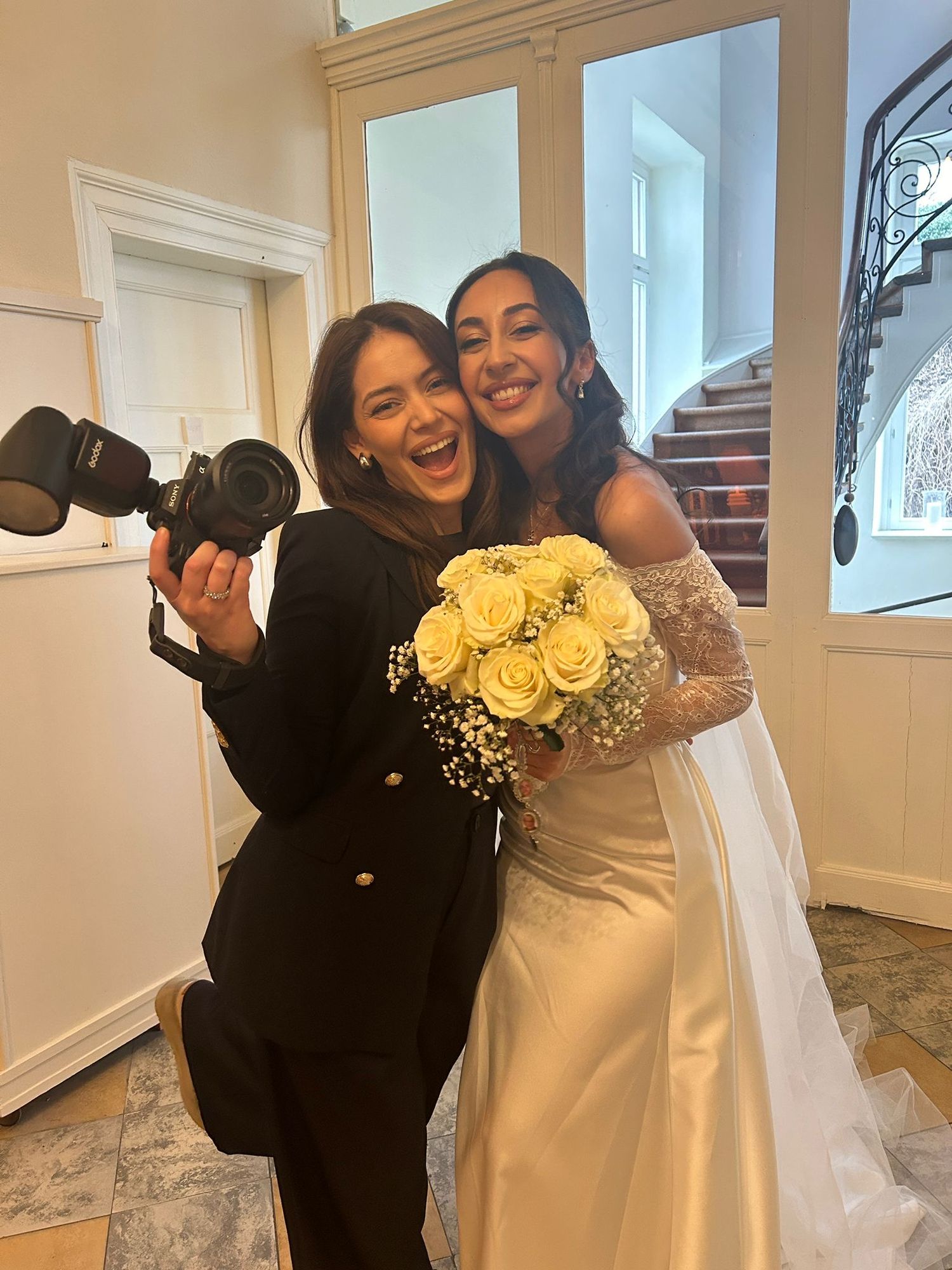 Photographer holding camera poses with bride in white gown holding bouquet of white roses indoors.