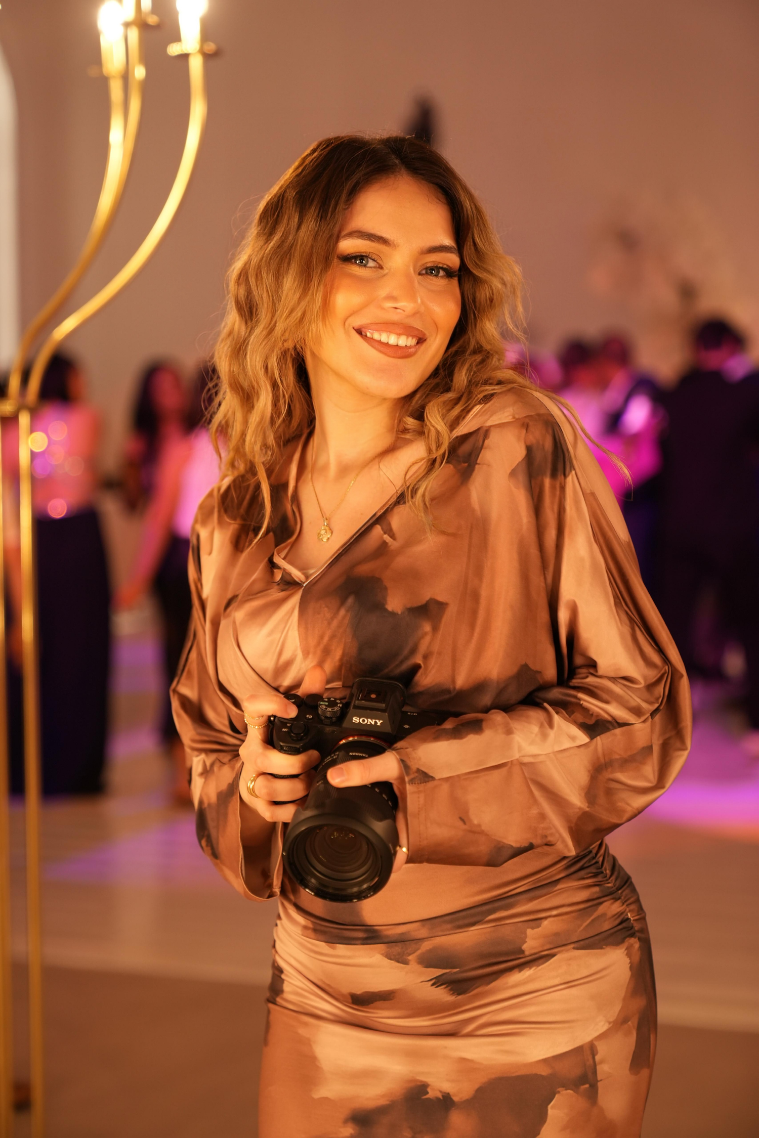 Smiling woman in satin outfit holds Sony camera at elegant event with purple lighting.