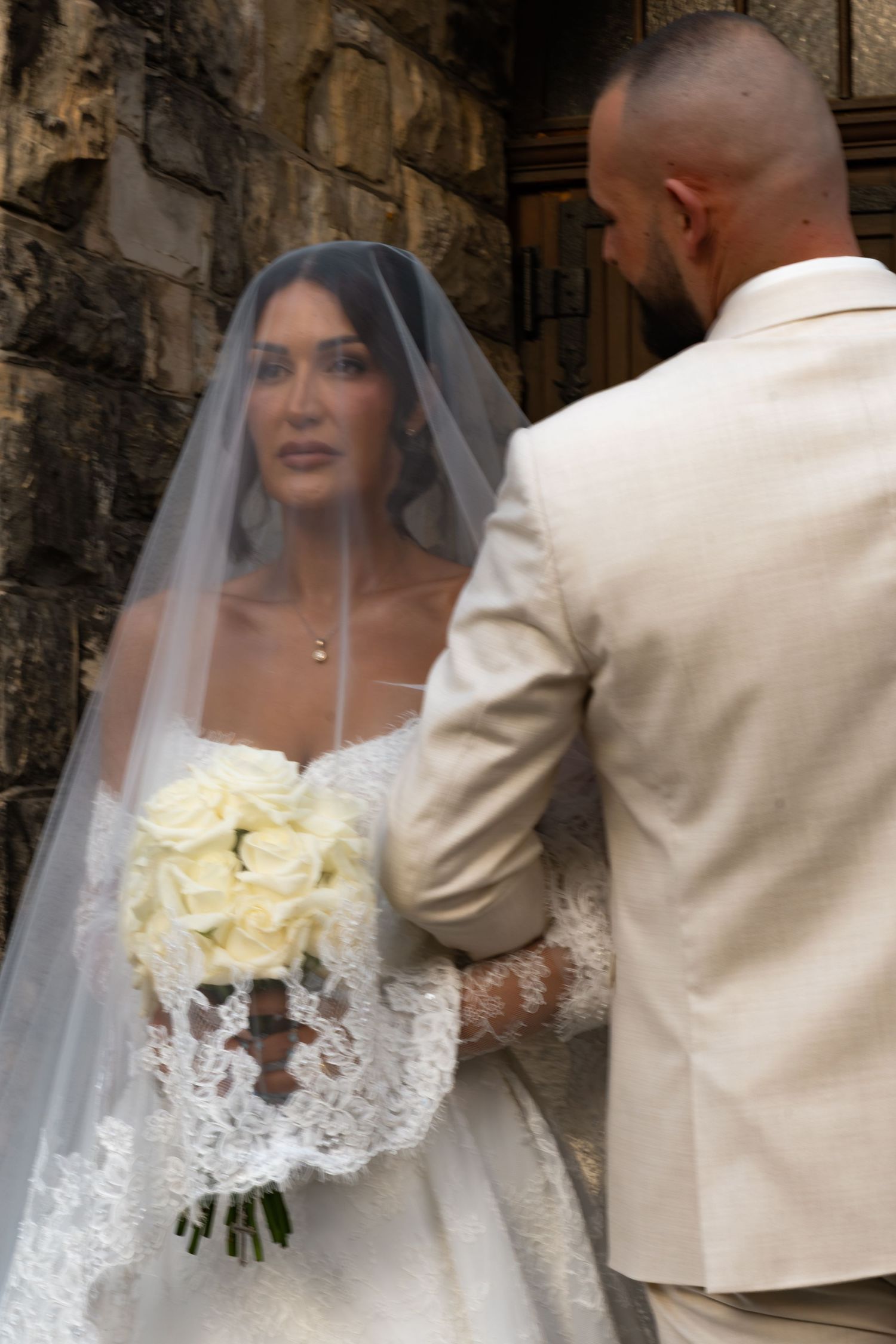 Bride in lace wedding dress and veil holds white rose bouquet facing groom in beige suit outside stone church.