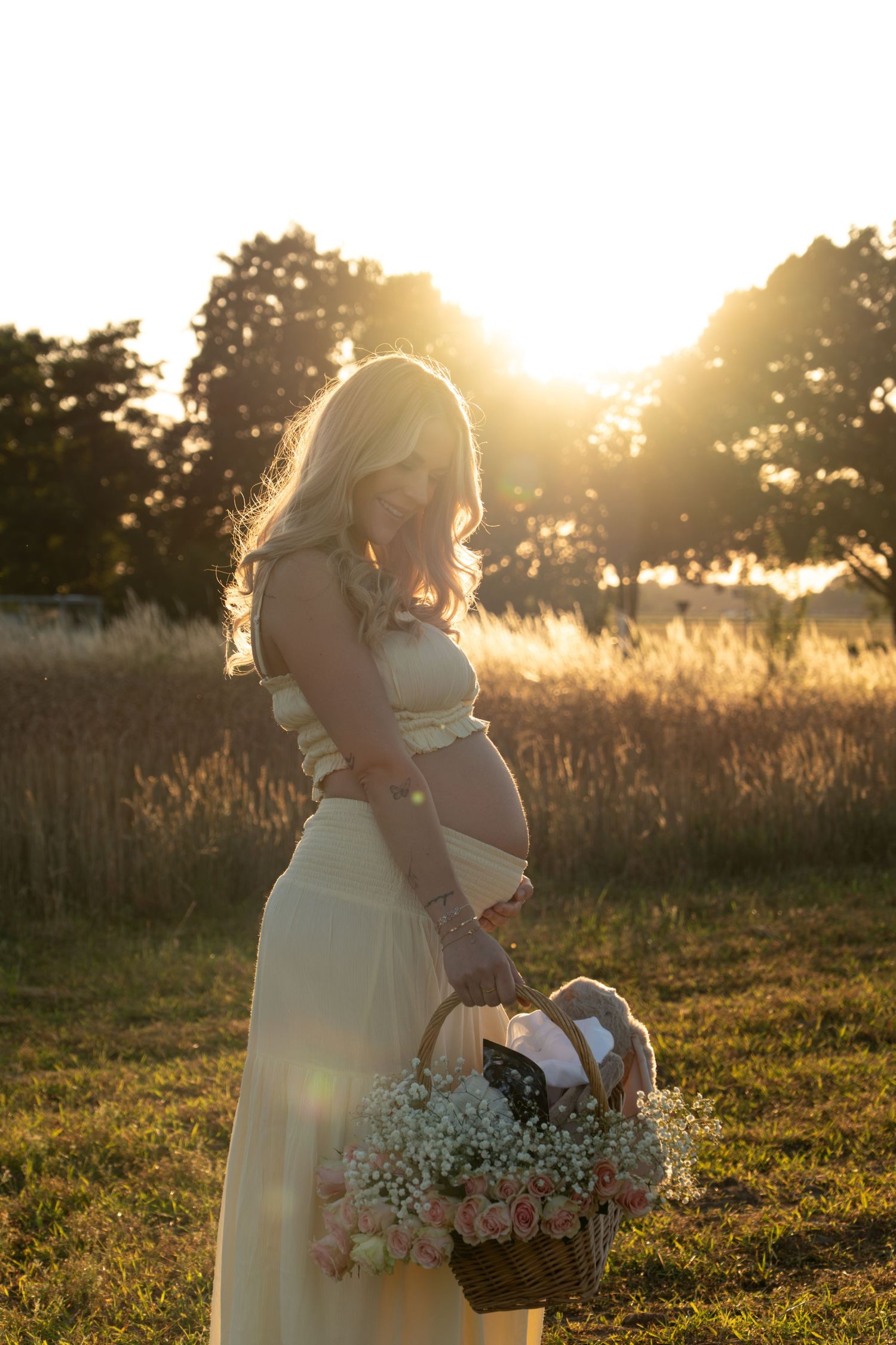 Pregnant woman in yellow dress cradling belly with flower basket in sunset field.