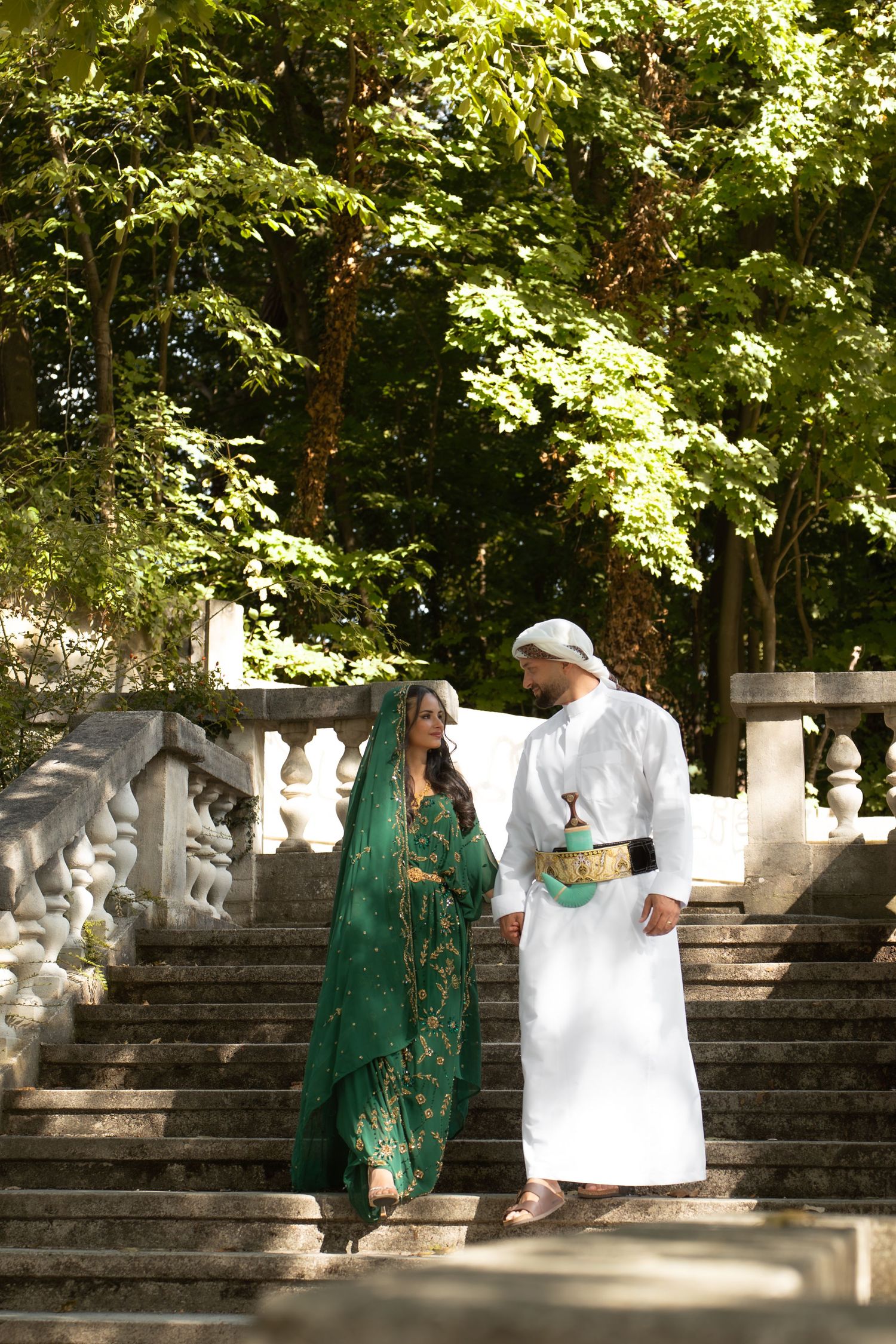 Couple in traditional Middle Eastern and Indian attire walking down ornate stone steps in a lush green park.