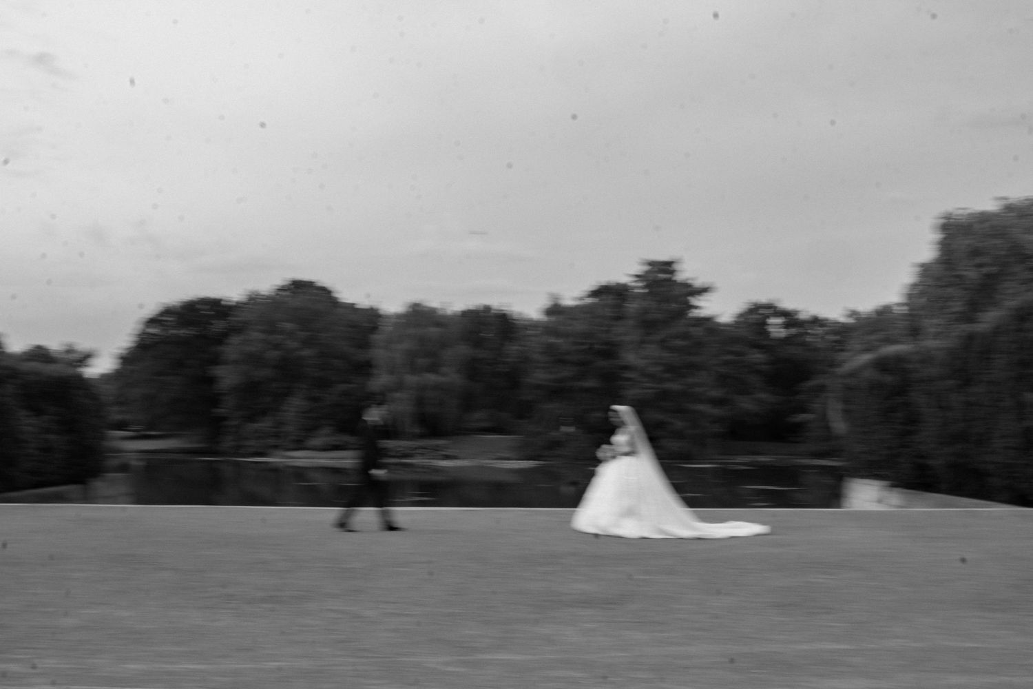 Black and white photo of bride in flowing gown walking across green field near lake.