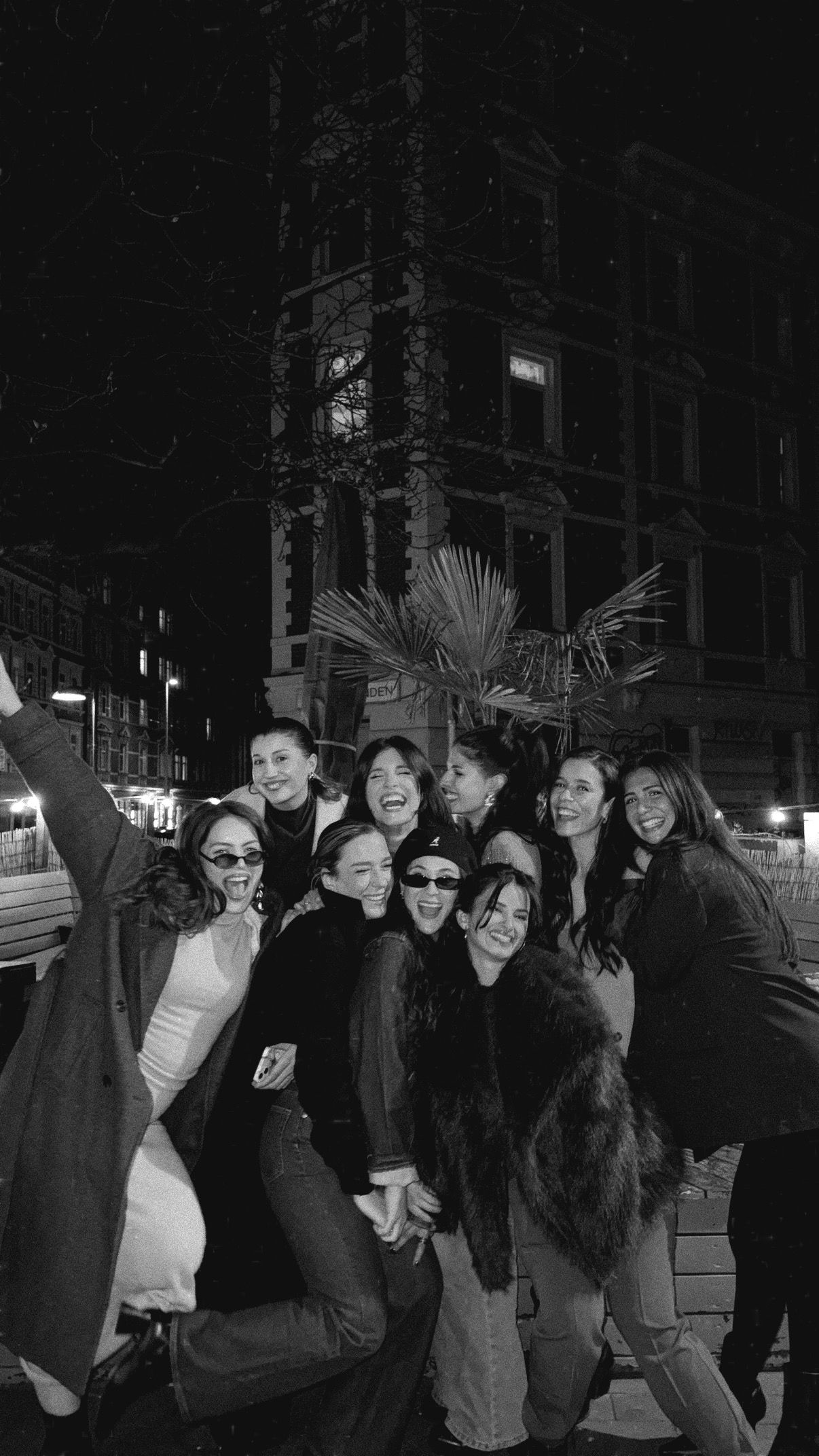 Black and white photo of a large group of women laughing and posing together on a city street at night.