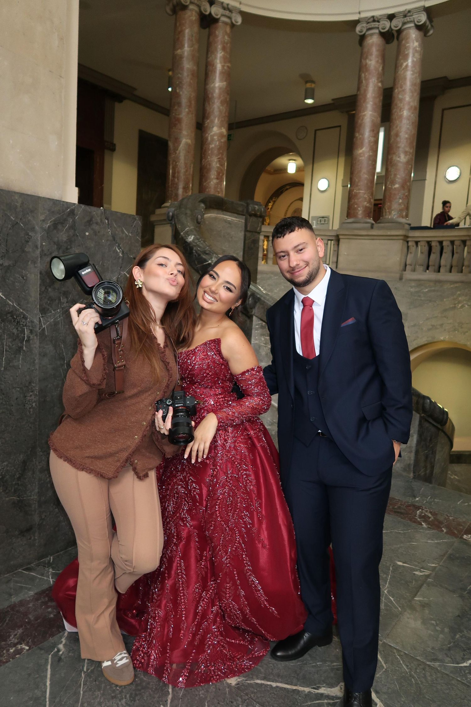 Female photographer poses with elegantly dressed couple in red gown and suit in grand hall.