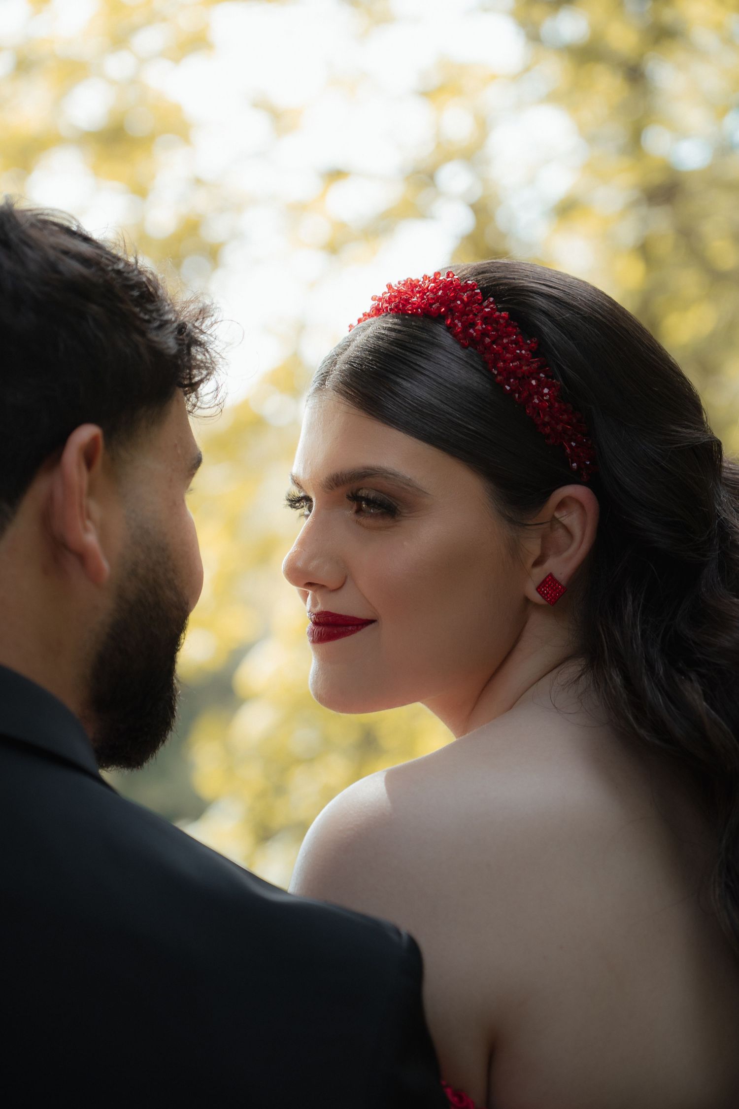 Close-up of bride with red headband and earrings gazing at groom in autumn outdoor setting.