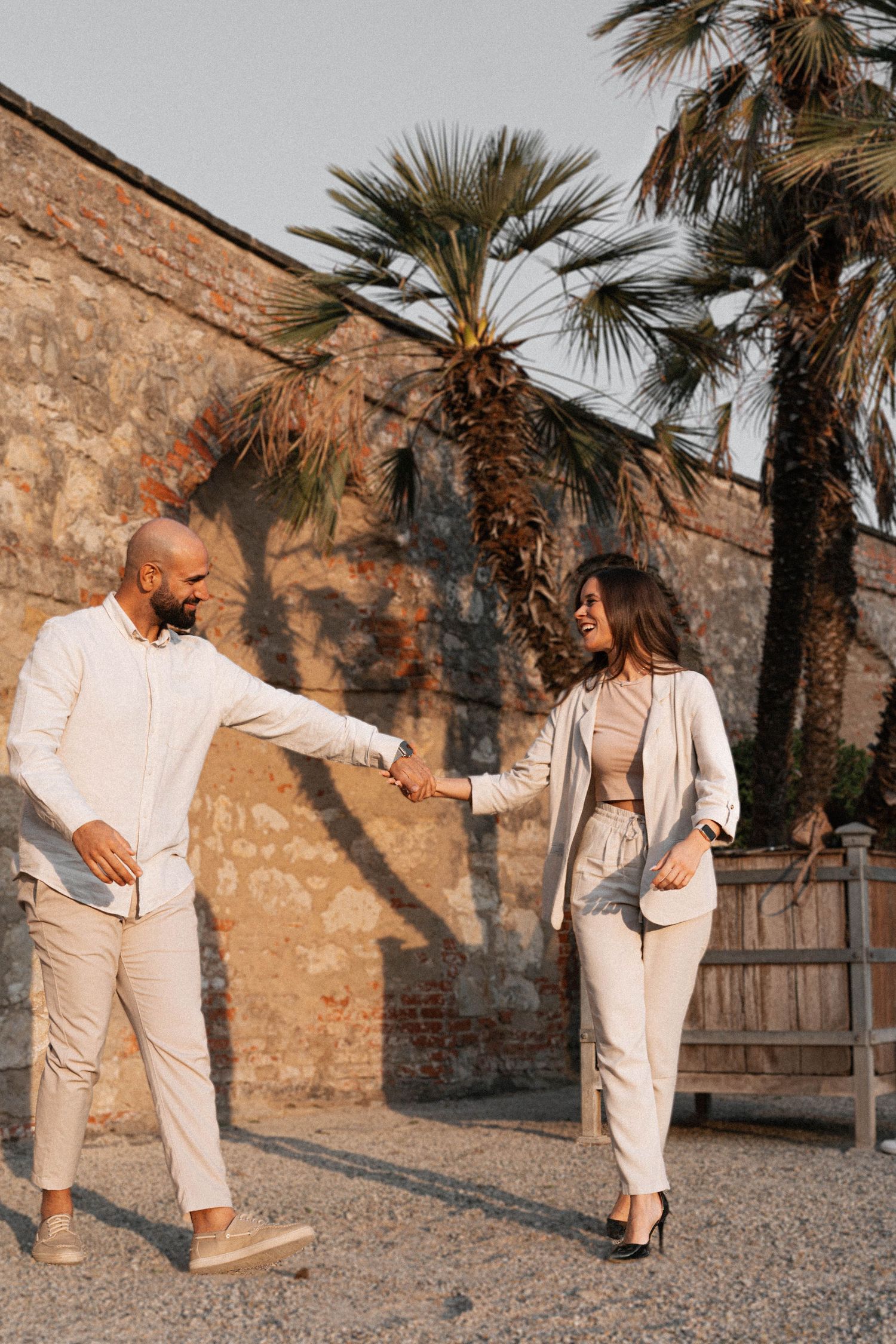 Couple in beige outfits holding hands and walking near palm trees and rustic stone wall.