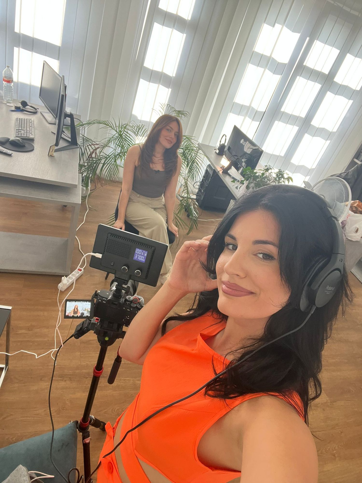 Woman in orange top with headphones takes selfie with camera on tripod in bright studio space.