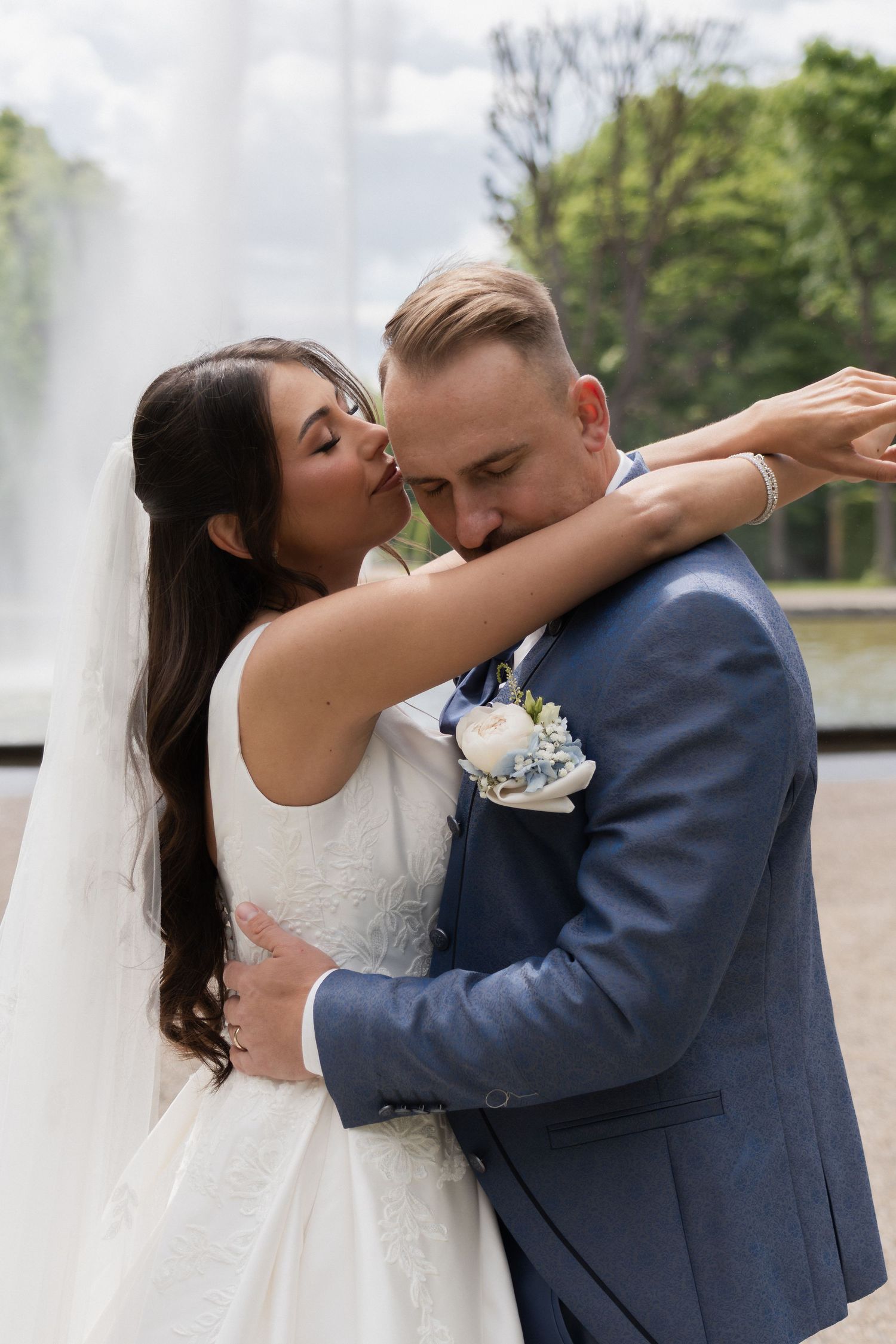 Bride kissing groom on cheek while embracing near fountain, groom wearing blue suit with boutonniere.