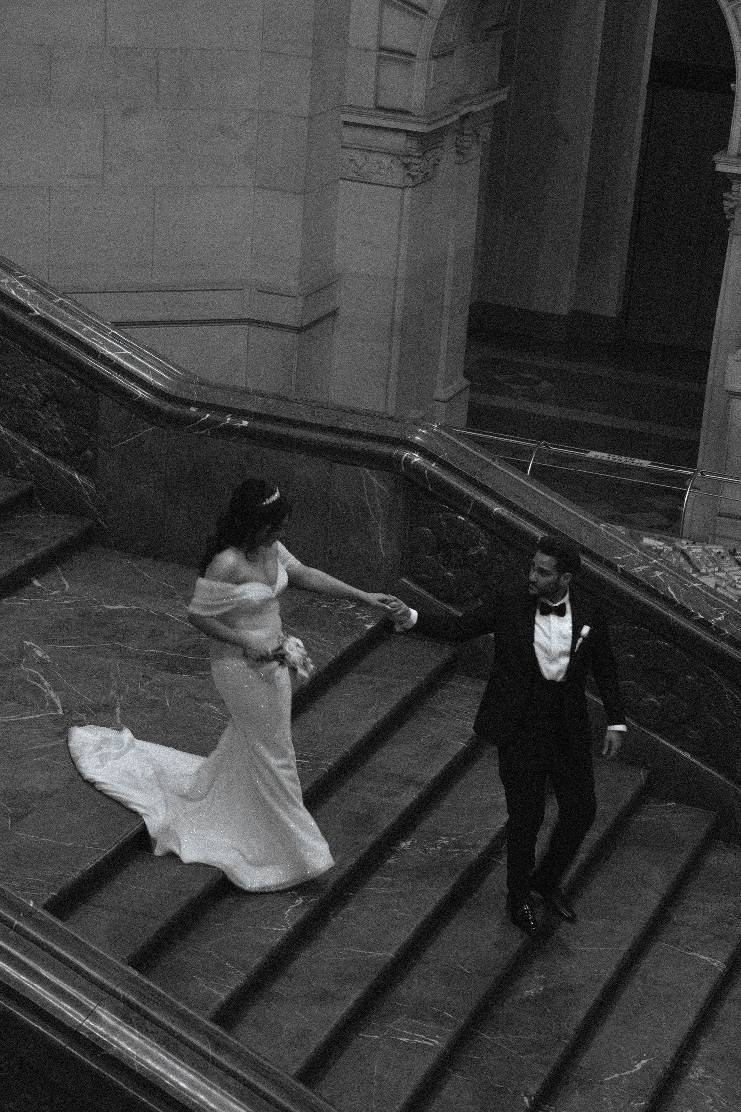 Black and white photo of bride and groom descending grand marble staircase in elegant wedding attire.