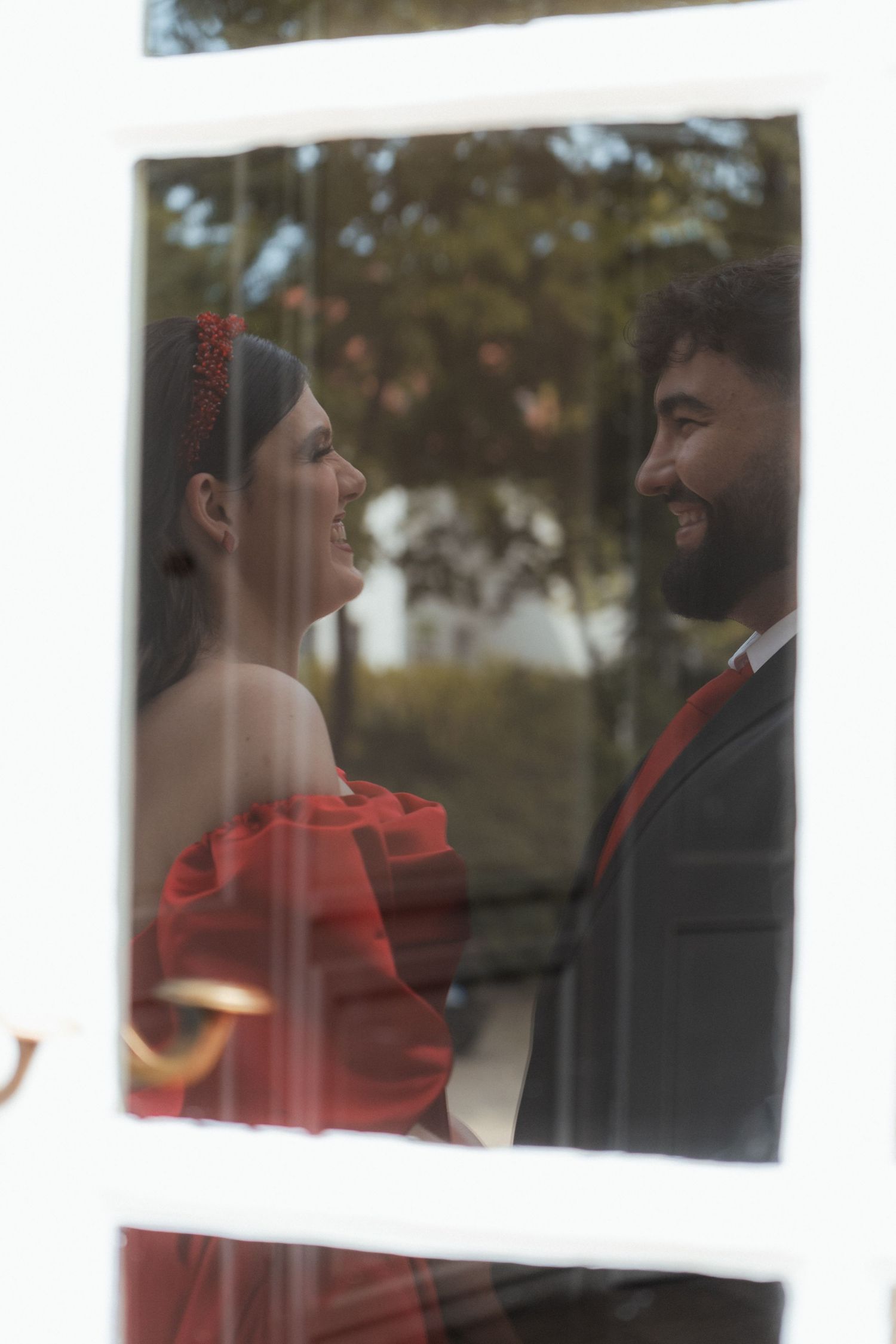 Couple in red dress and suit smiling at each other through a window with trees in background.