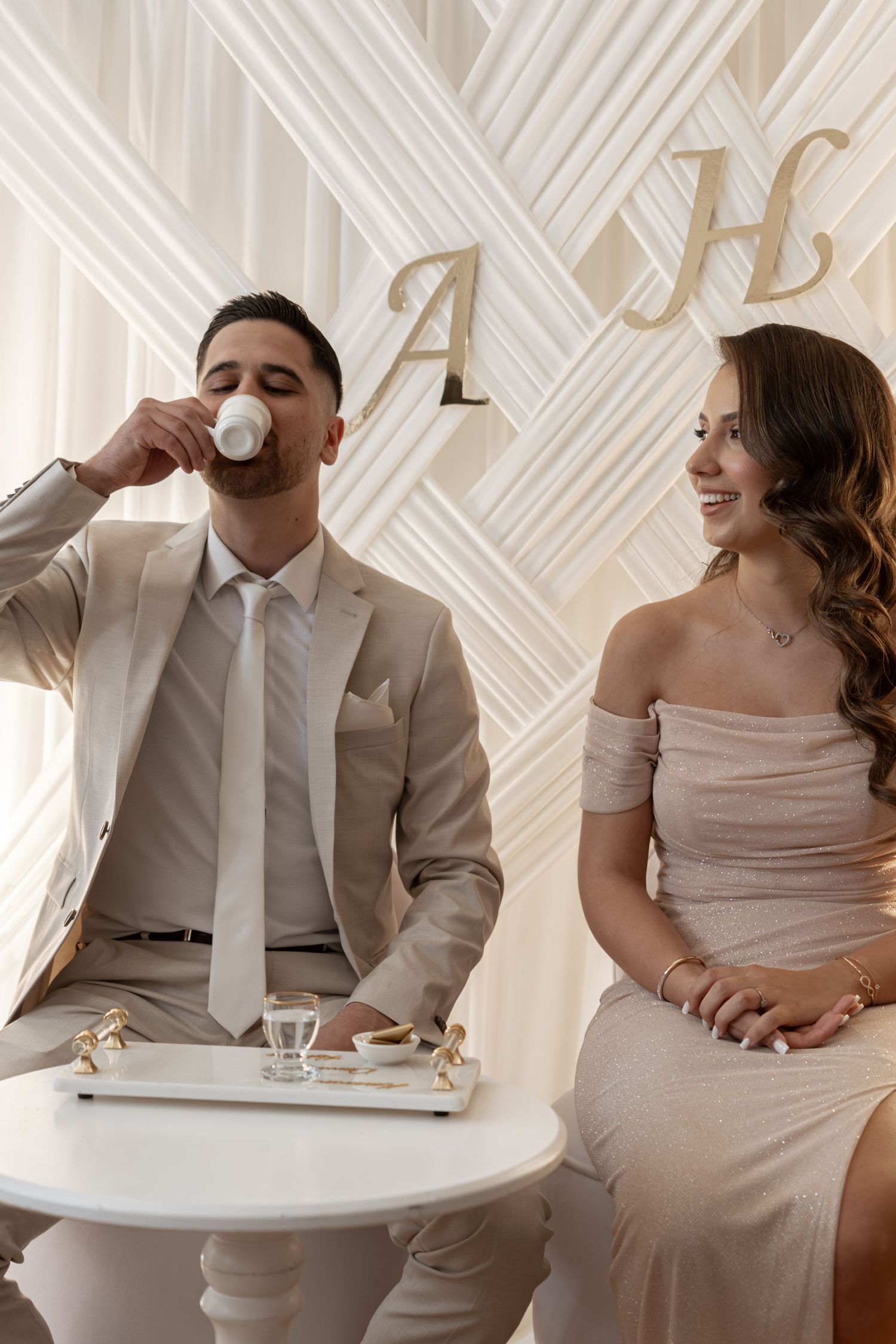 Couple in beige outfits sitting at white table in front of decorative monogram backdrop.
