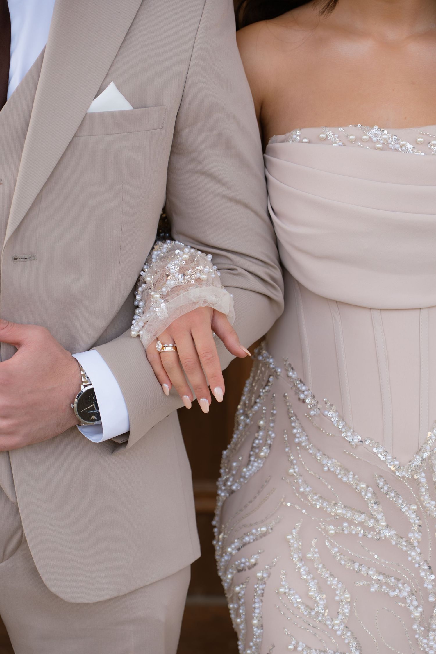 Close-up of bride and groom in beige embellished outfits showing wedding rings and watch.