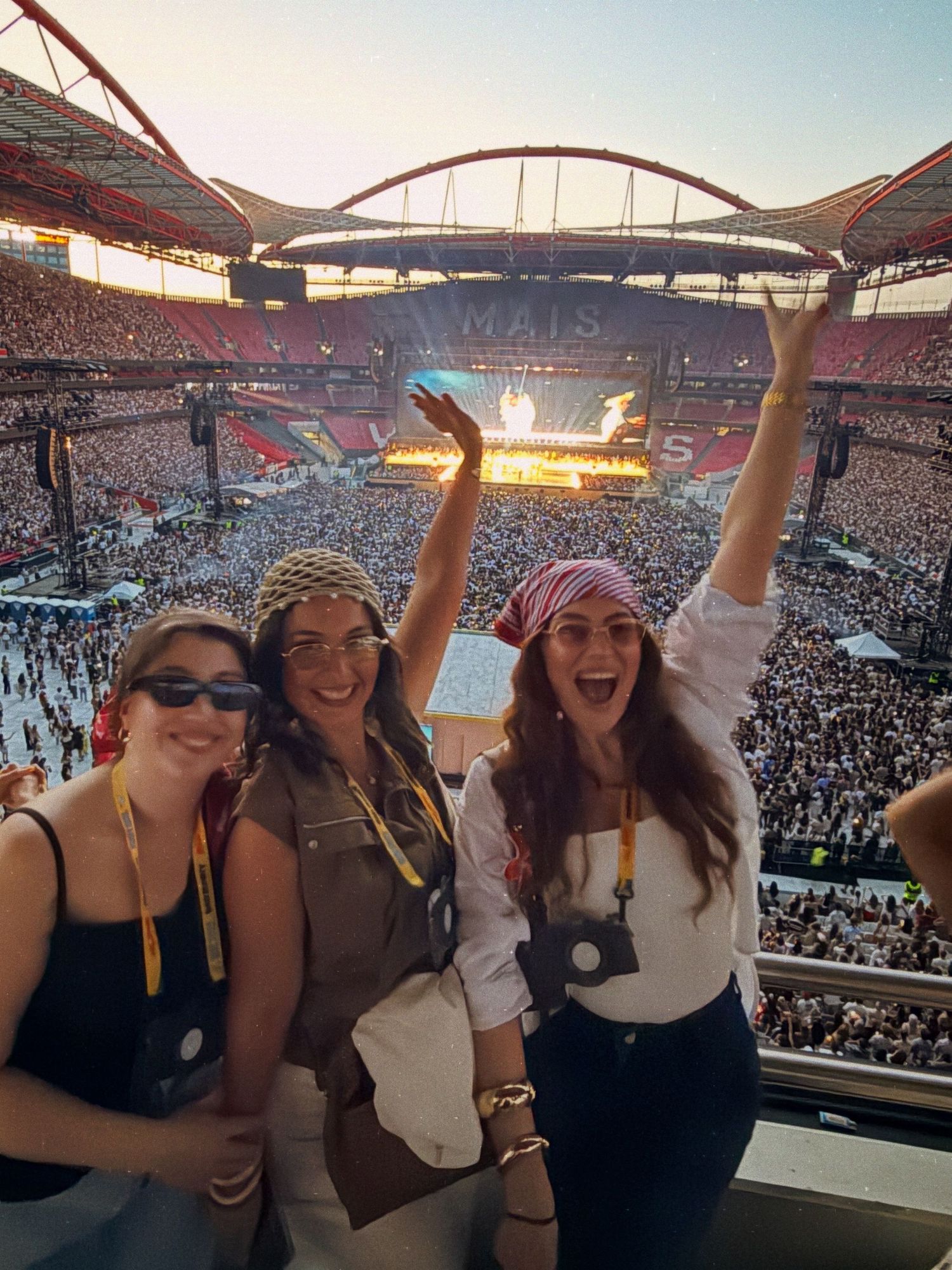 Three women smiling at a packed concert stadium at sunset, one wearing a red bandana headscarf.