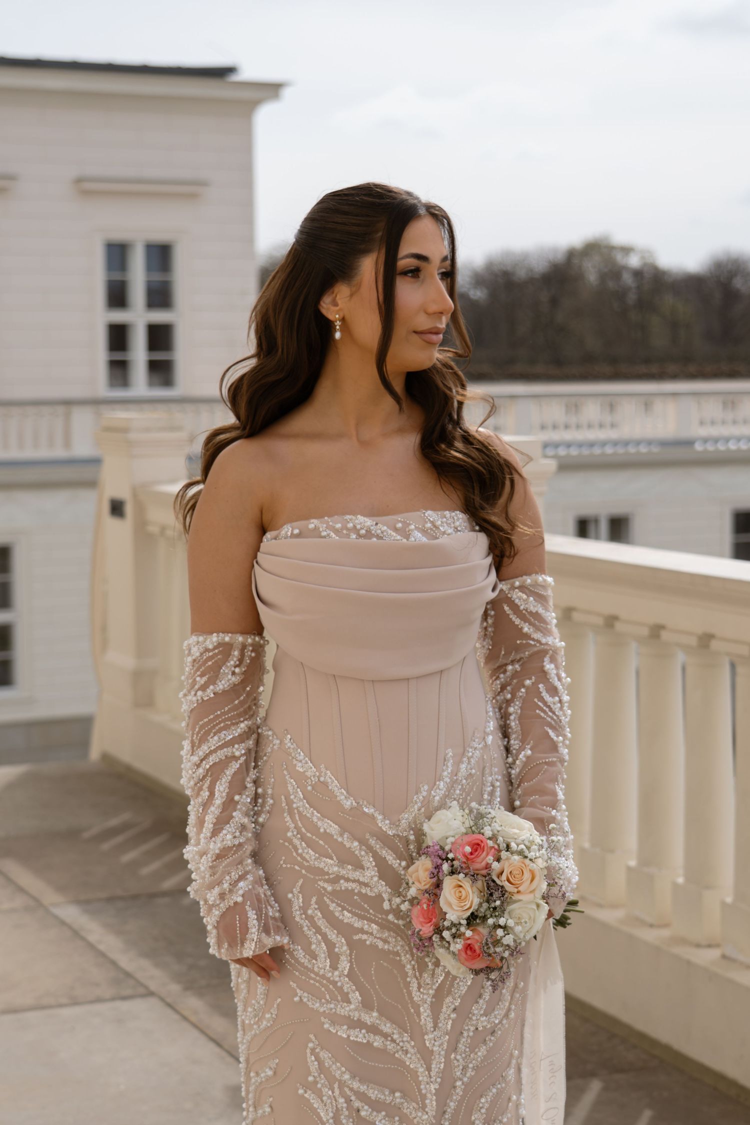 Bride in strapless beige lace gown with long sleeves holding bouquet on outdoor balcony.