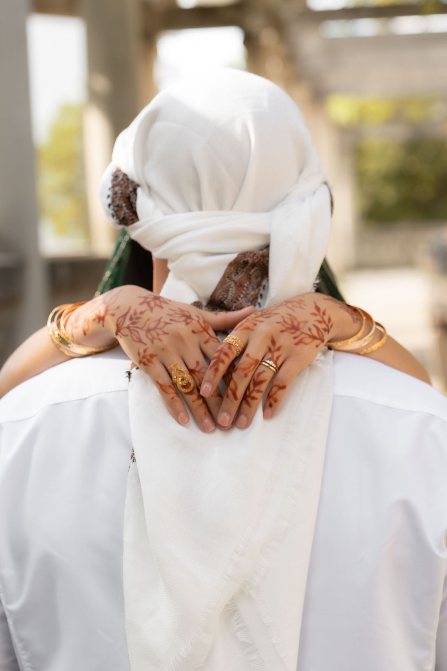 South Asian bride with intricate henna hands and gold bangles embraces groom from behind at outdoor wedding ceremony.