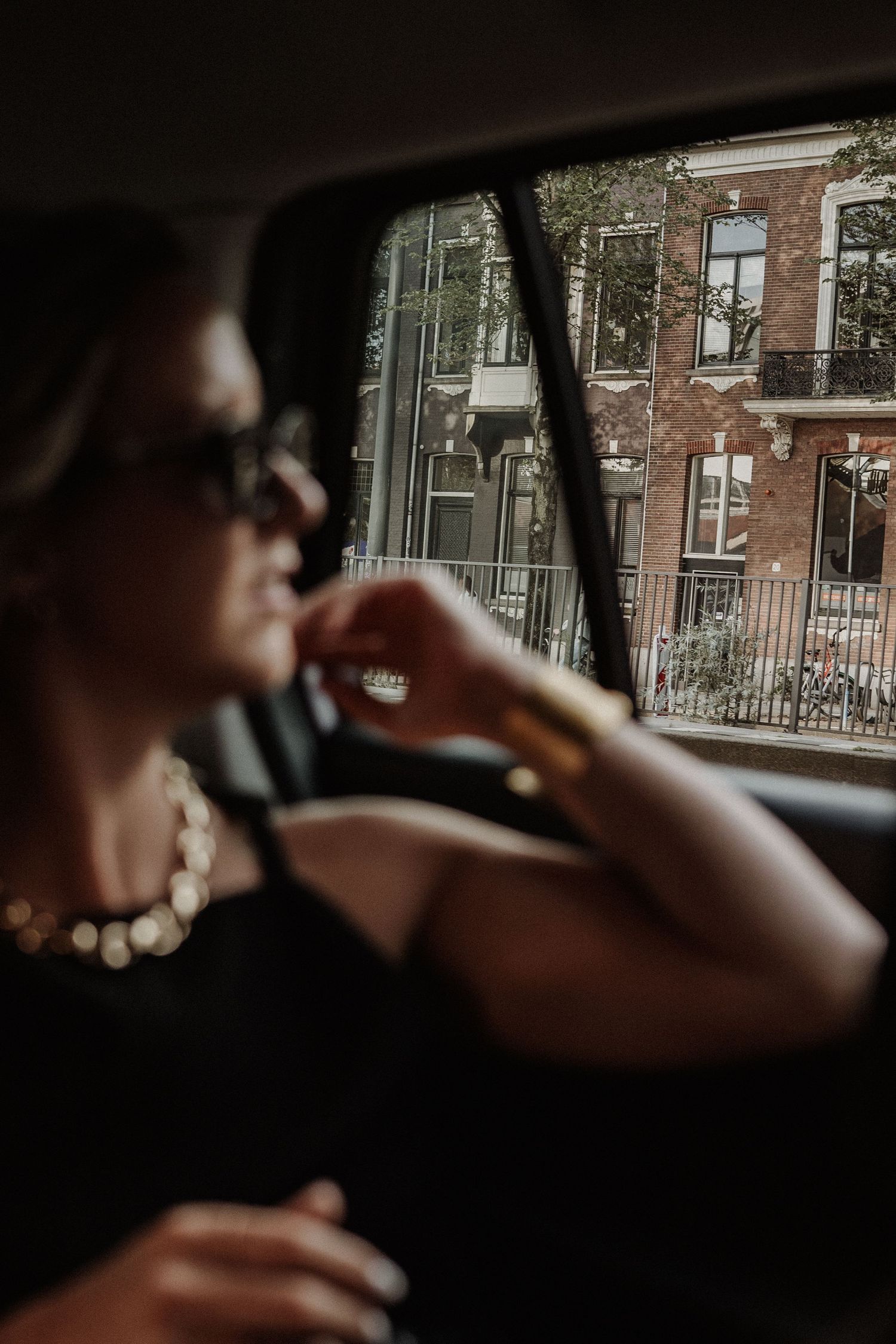 A silhouette sits in a car with pearl necklace and sunglasses, while brick buildings are visible through the window.