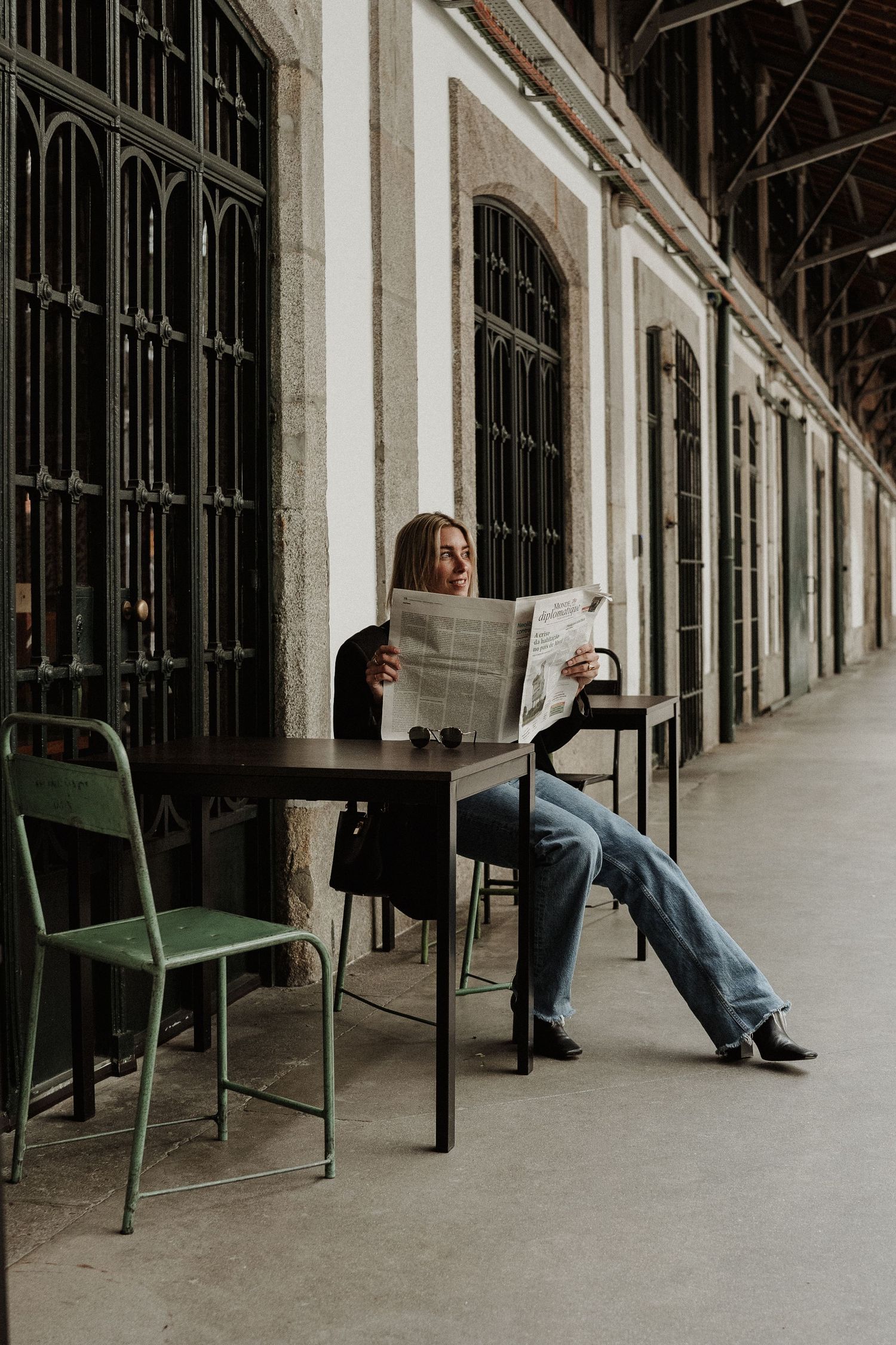 A person sits casually reading a newspaper at a small table in a covered colonnade with arched windows.