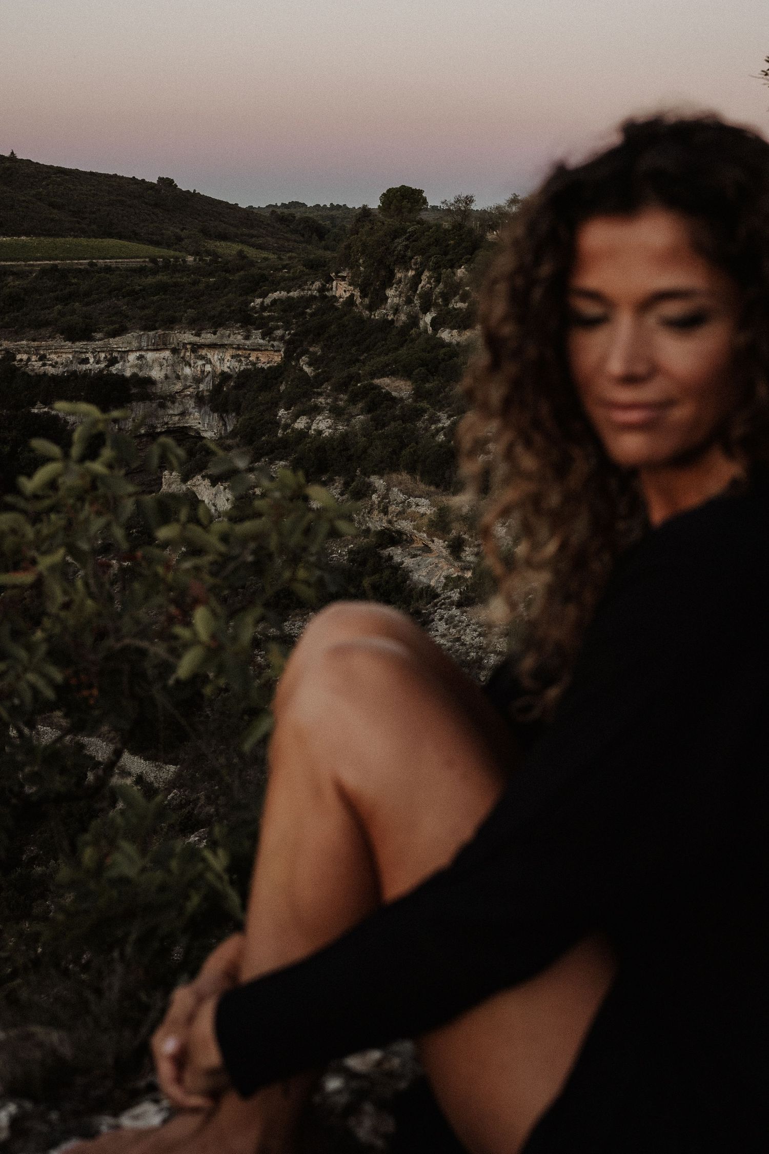 Person with curly hair in a black sweater sits outdoors at sunset against a scenic cliff landscape.