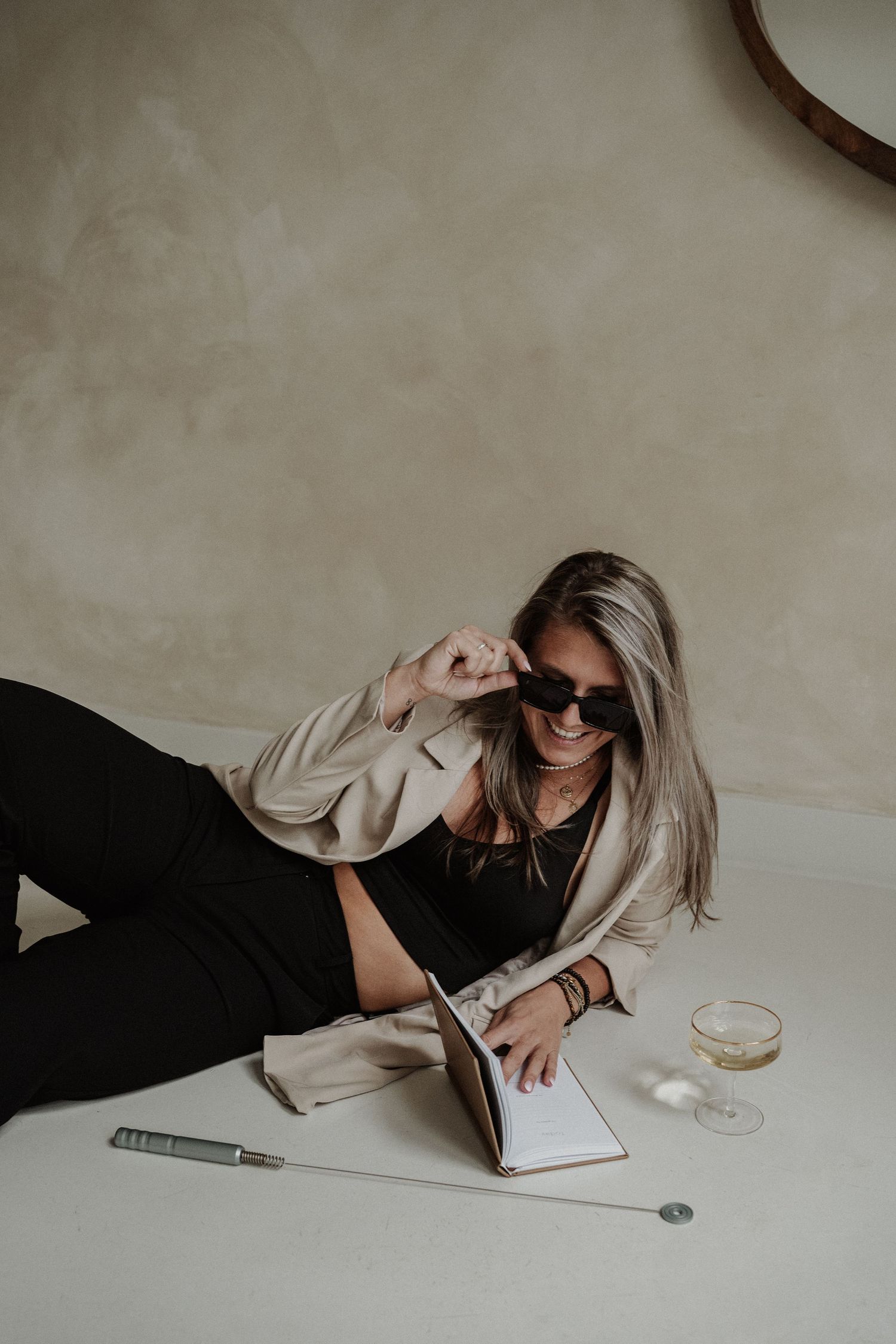 Woman in sunglasses lounges on floor while writing in notebook with coffee cup nearby, wearing casual beige and black outfit.