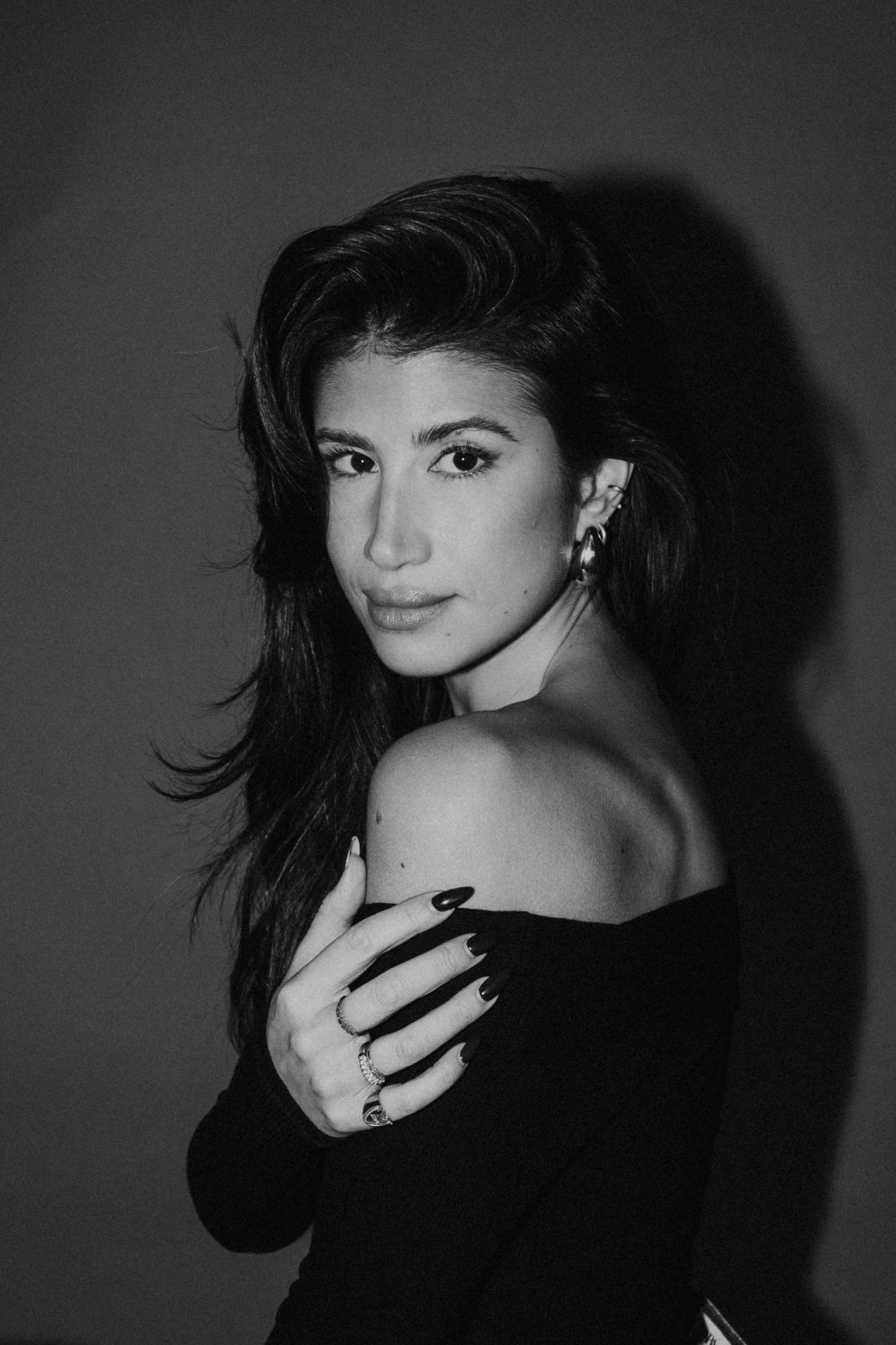 Black and white portrait of a woman in an off-shoulder black top looking over her shoulder with dark flowing hair.