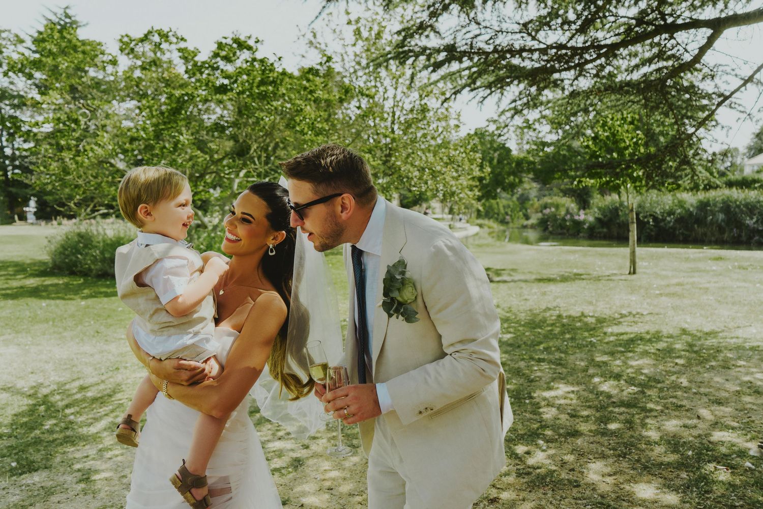 Happy bride and groom walking outdoors holding baby in sunny garden.