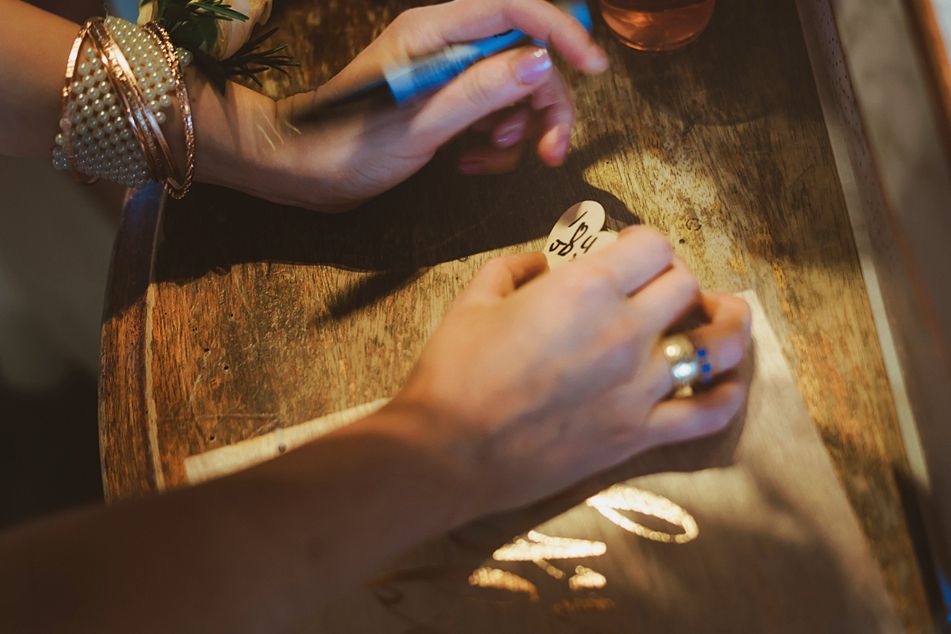 Close-up of hands writing on small stones at a rustic wooden table, adorned with bracelets and rings.
