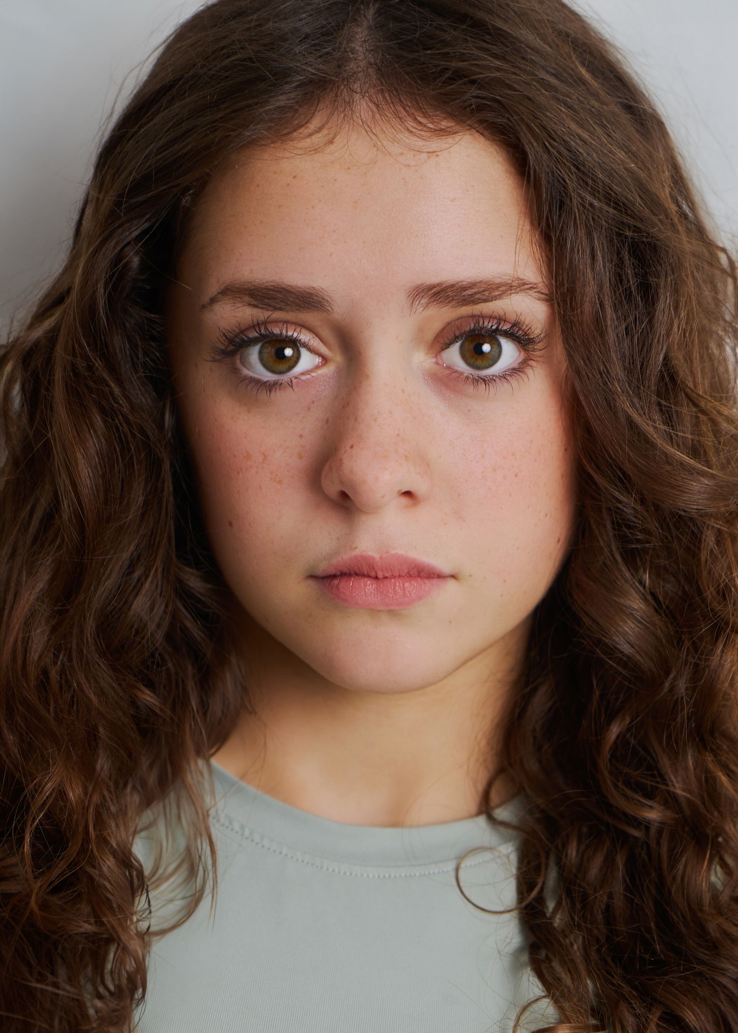 Close-up portrait of a young woman with curly brown hair, freckles, and hazel eyes in a light blue top.