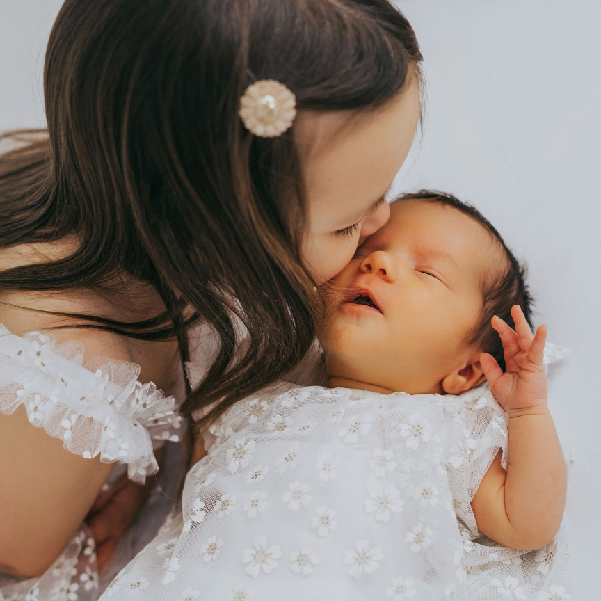 Young girl in white lace dress gently kissing a sleeping newborn baby on a soft white background.