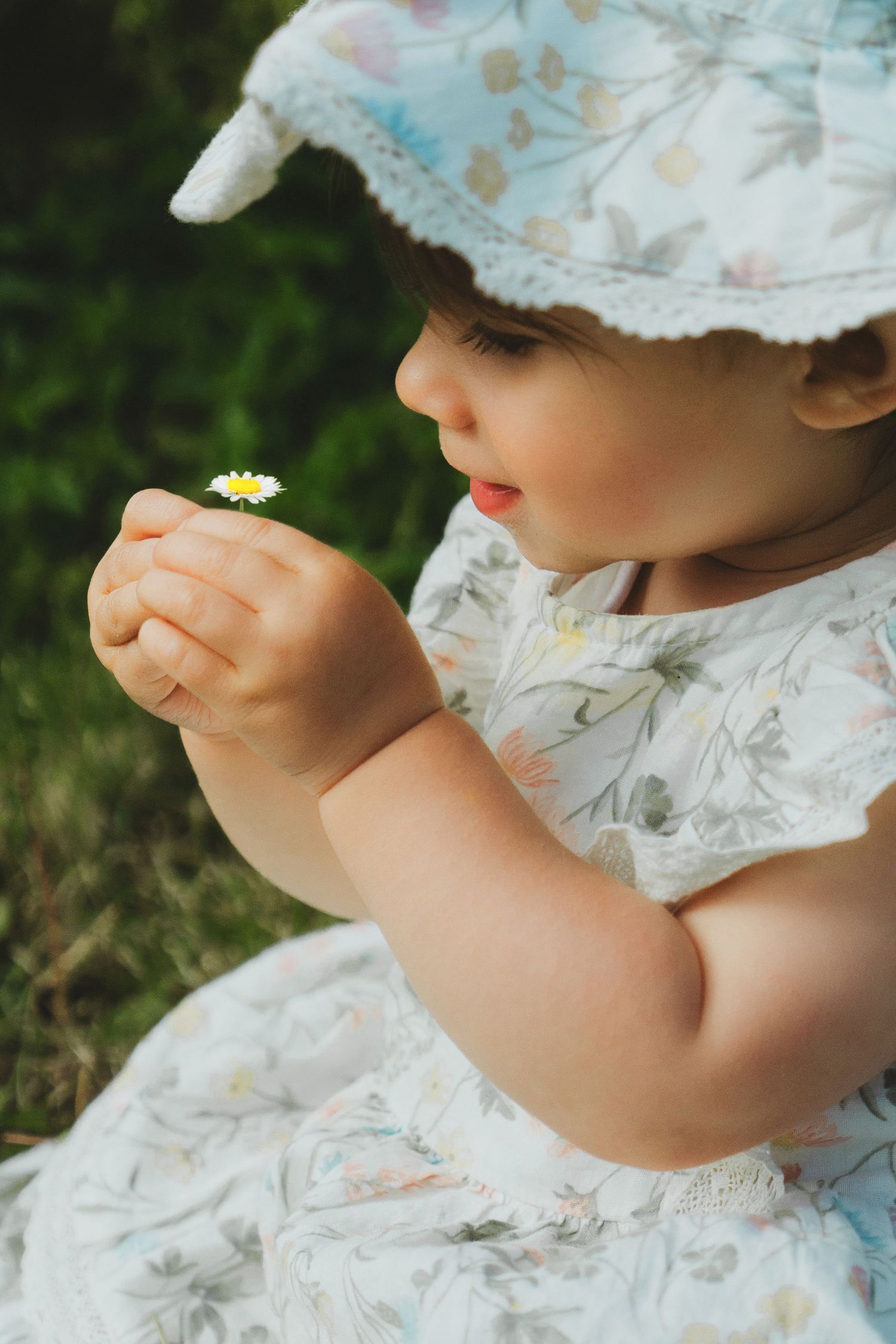 Baby girl in floral dress and hat holds a tiny daisy flower outdoors in green grass.