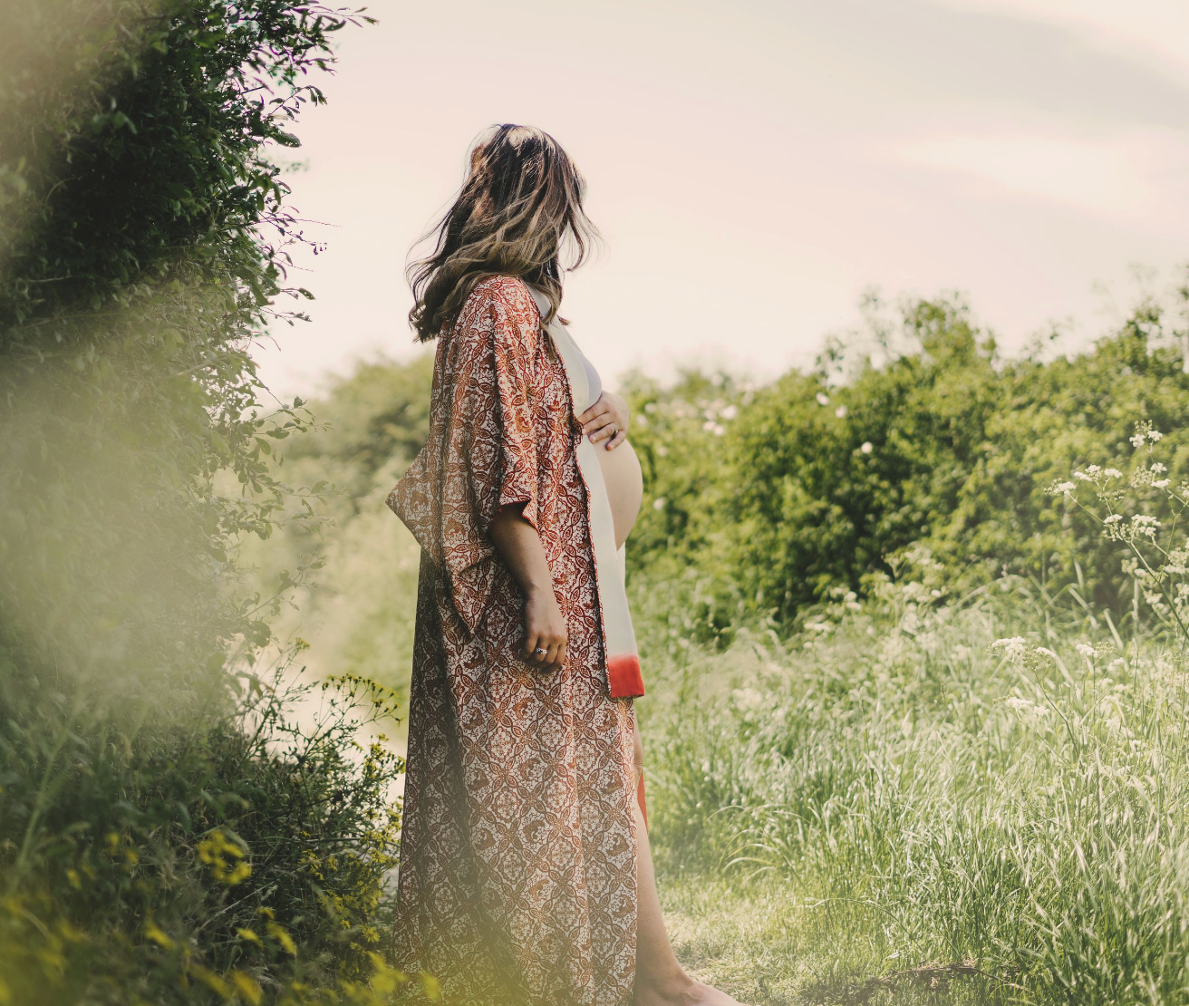 Pregnant woman in floral maxi dress standing in lush green meadow, cradling baby bump on sunny day.