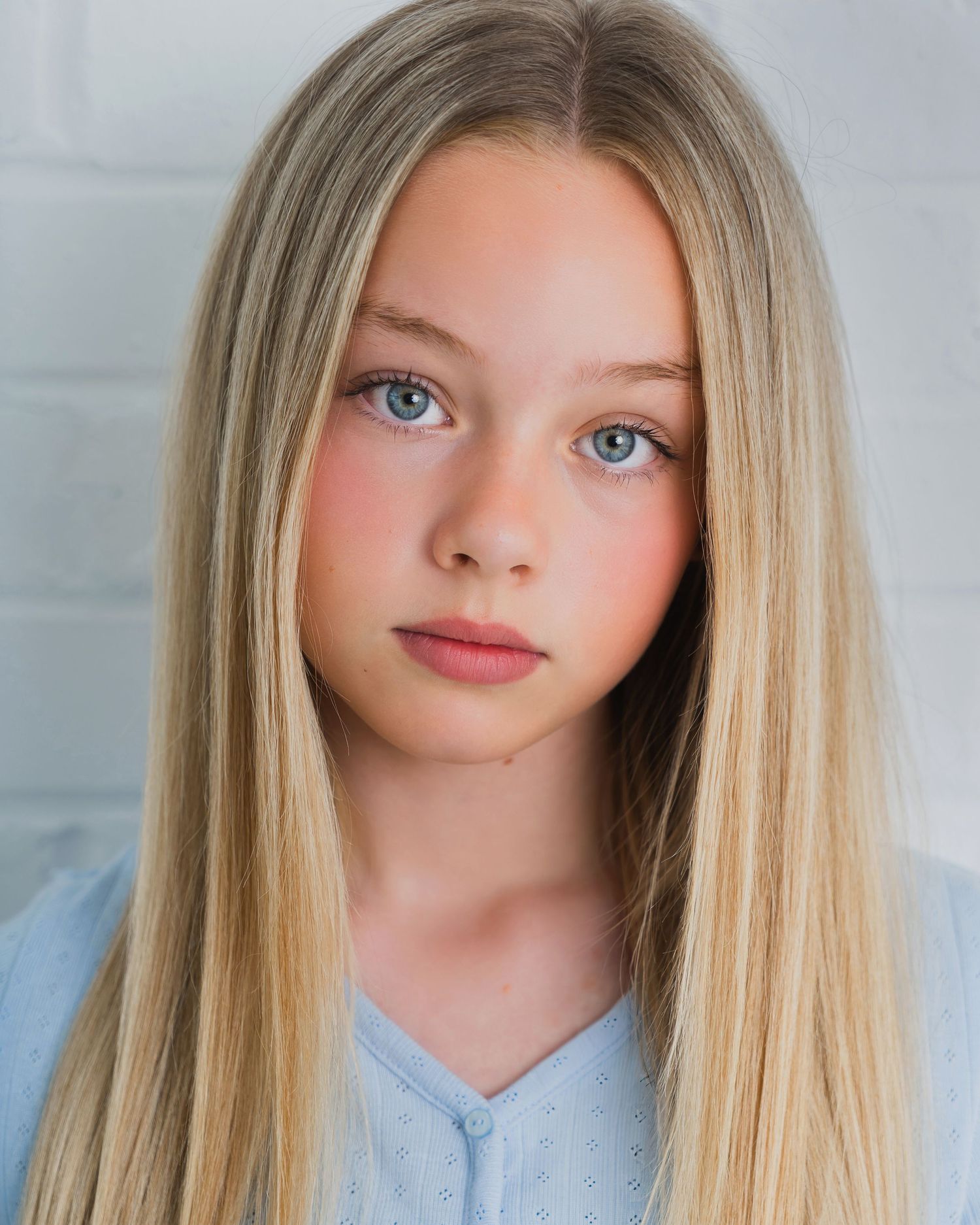 Young blonde girl with blue eyes and long straight hair wearing a light blue top against white background.