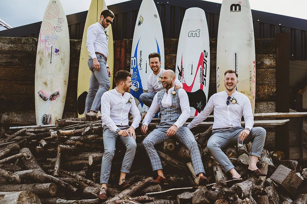 Groom and groomsmen laughing beside colorful surfboards in casual wedding photo.