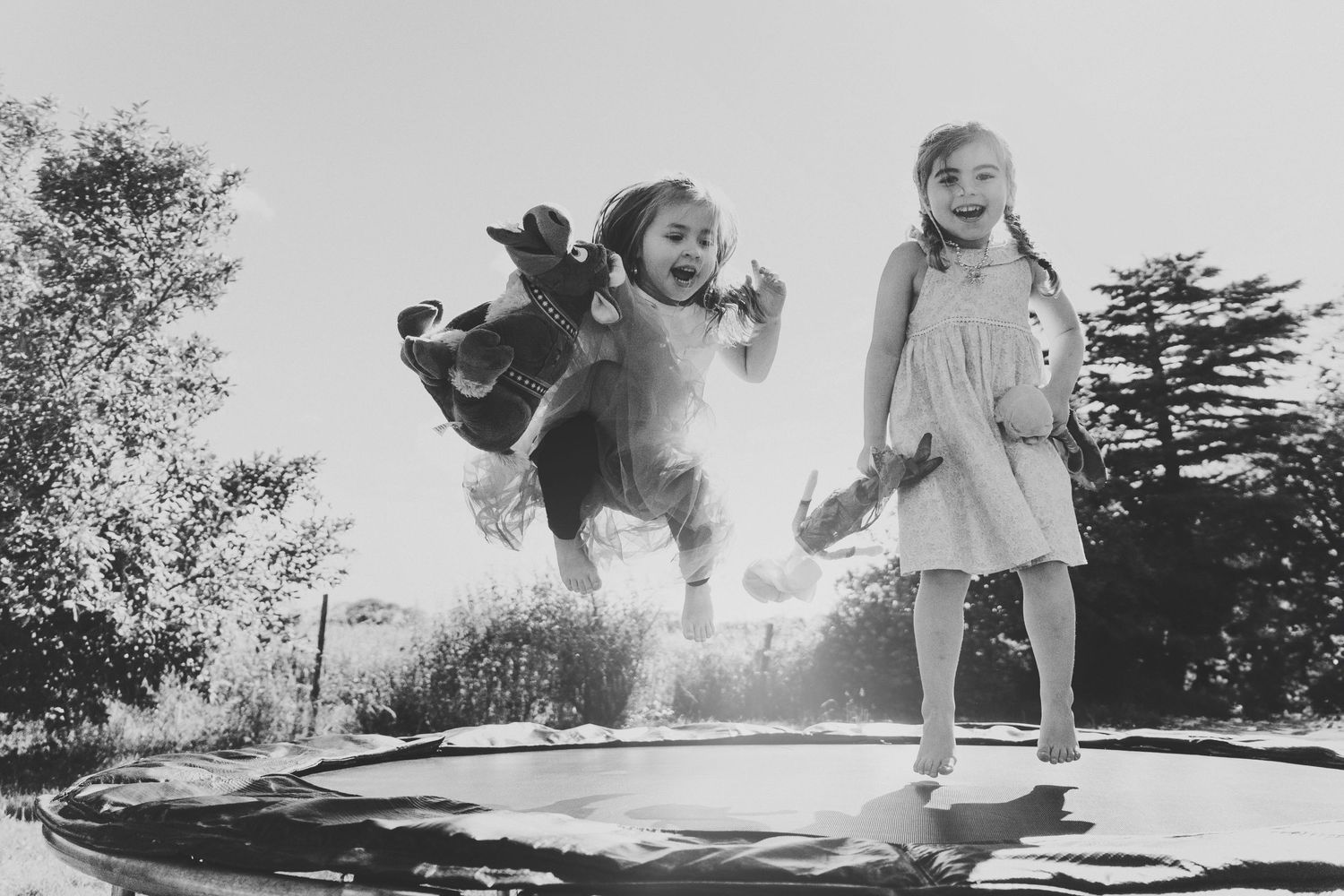 Black and white photo of two girls jumping joyfully on a trampoline in a sunny backyard.